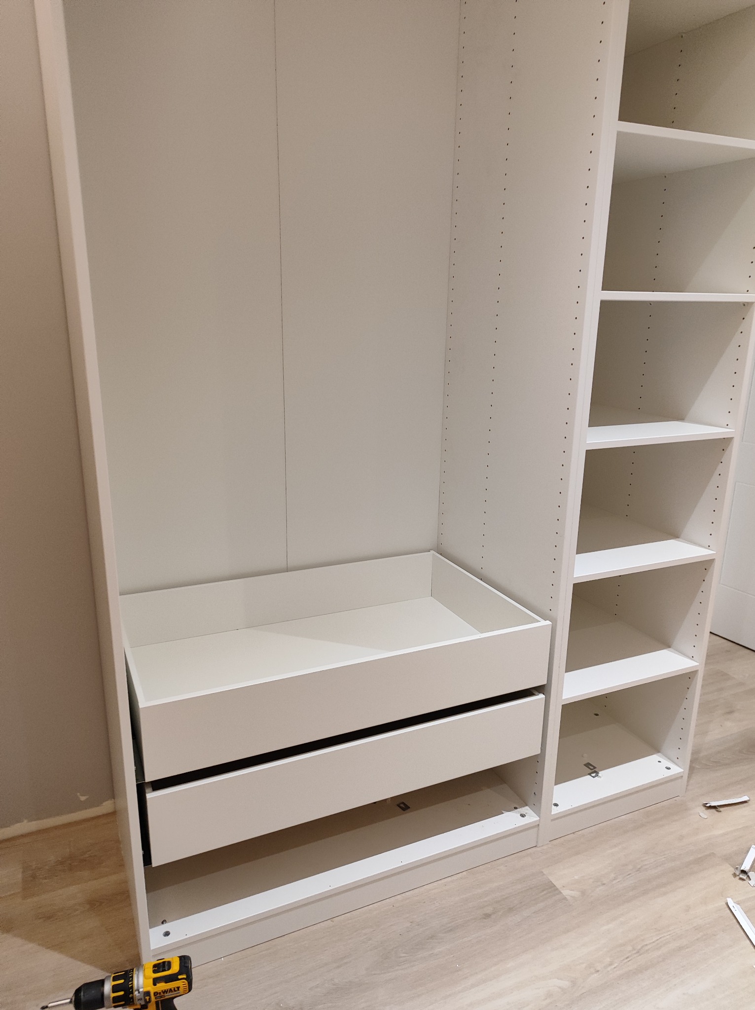 Biała, modułowa szafa z szufladami i półkami w trakcie montażu, widoczna wkrętarka DeWalt na podłodze.