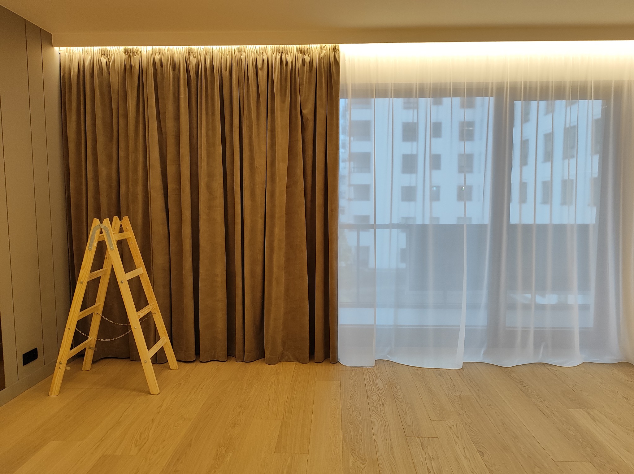 Montaż zasłon: drewniana drabina oparta o ścianę z brązowymi, aksamitnymi zasłonami, obok okna z białymi, prześwitującymi firankami, widoczny parkiet na podłodze, oświetlenie LED podsufitowe.