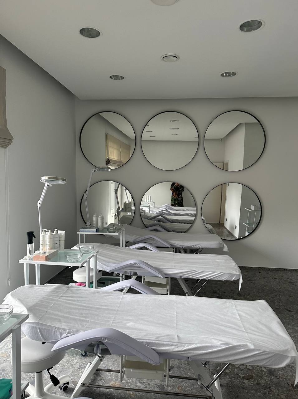 Jasne wnętrze gabinetu kosmetycznego z trzema przygotowanymi stanowiskami zabiegowymi, białymi leżankami i okrągłymi lustrami na ścianie, w których odbija się osoba robiąca zdjęcie.