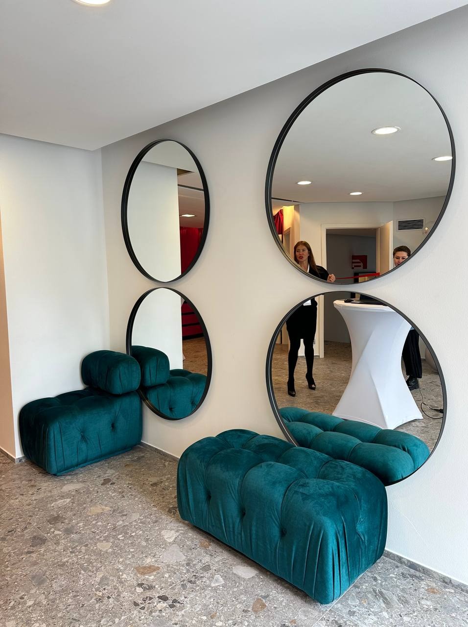 Nowoczesna aranżacja wnętrza z czterema okrągłymi lustrami w czarnych ramach na białej ścianie, odbijającymi fragmenty pomieszczenia i dwie osoby, oraz dwoma turkusowymi pufami o pikowanej fakturze...