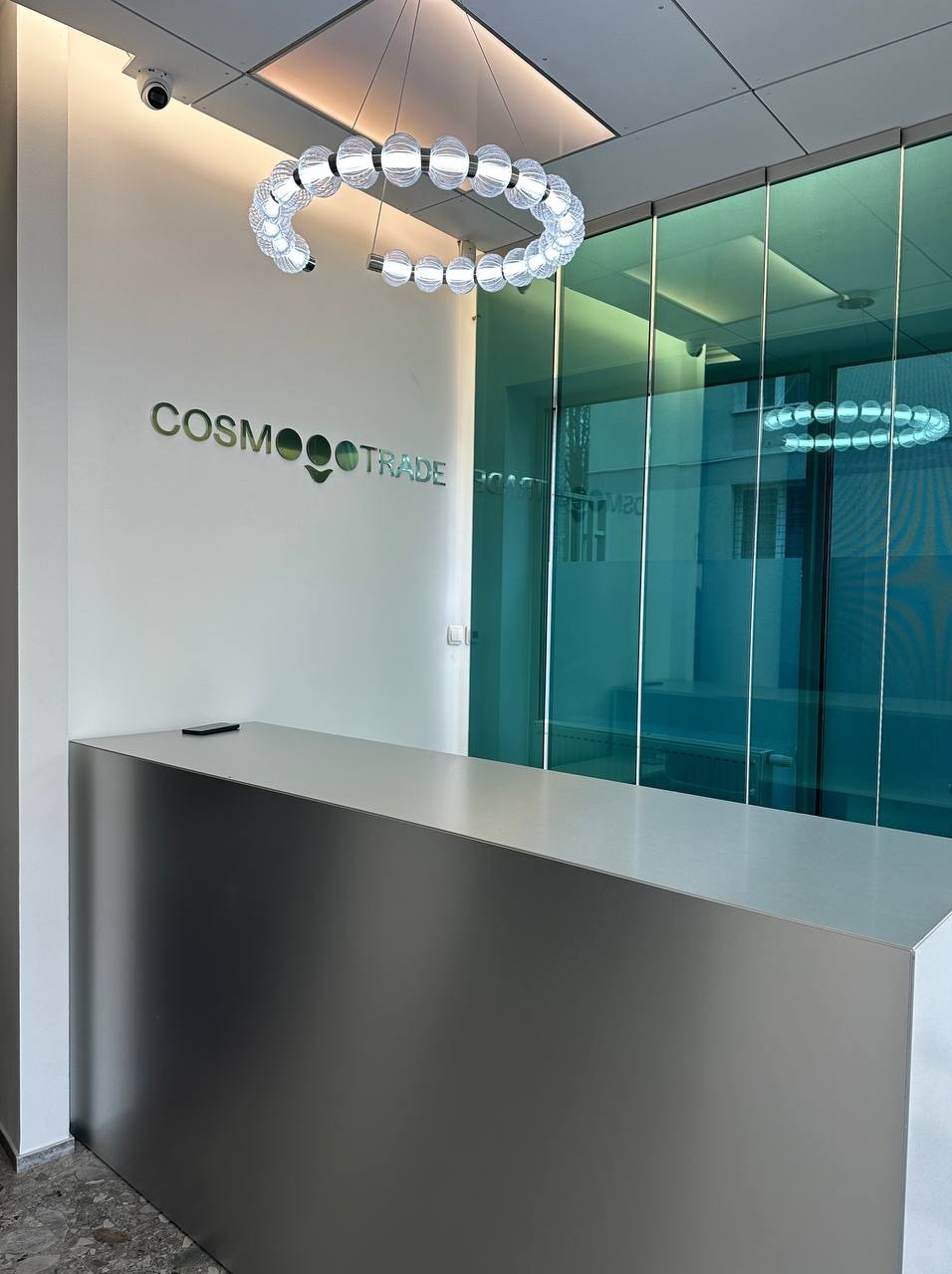 Nowoczesna recepcja biurowa z metalowym blatem, szklanymi ścianami działowymi i designerską lampą sufitową w kształcie pierścienia, logo firmy 'Cosmo Trade' na ścianie.
