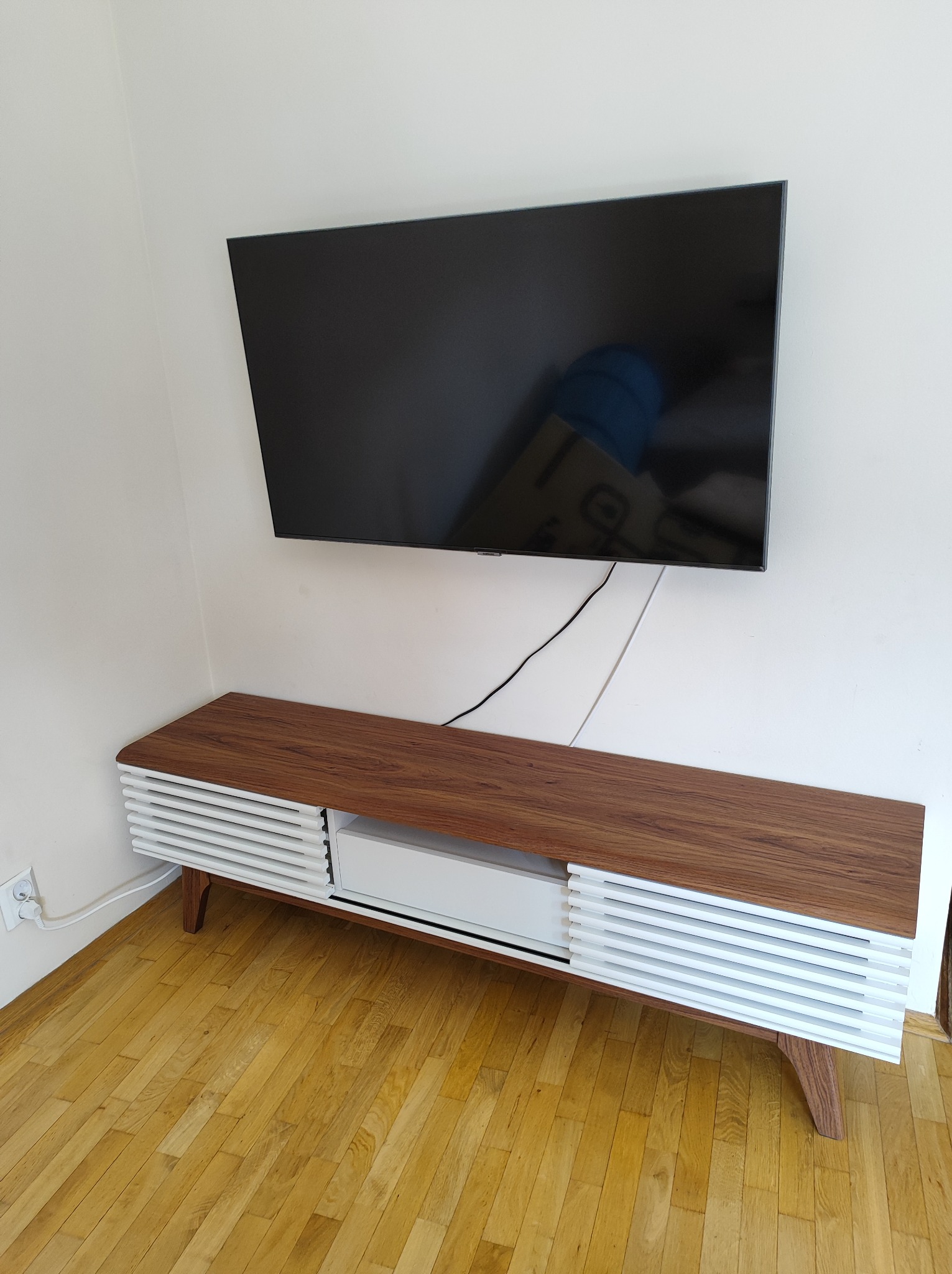 Nowoczesna szafka RTV z blatem w kolorze orzecha i białymi, lamelowymi frontami, umieszczona pod wiszącym telewizorem na białej ścianie, widoczna na tle parkietu.