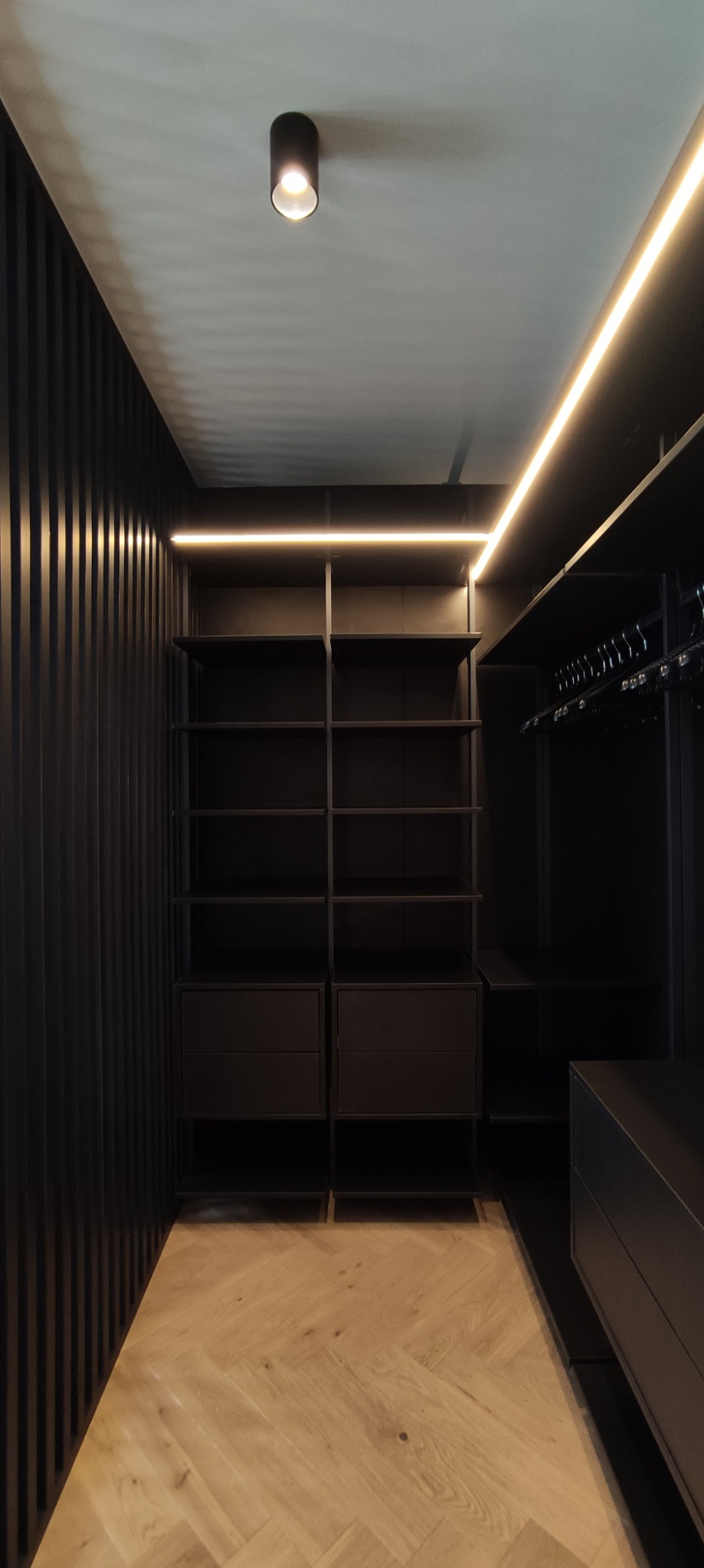Ciemna garderoba z oświetleniem LED, drewnianą podłogą w jodełkę i czarnymi lamelami na ścianie