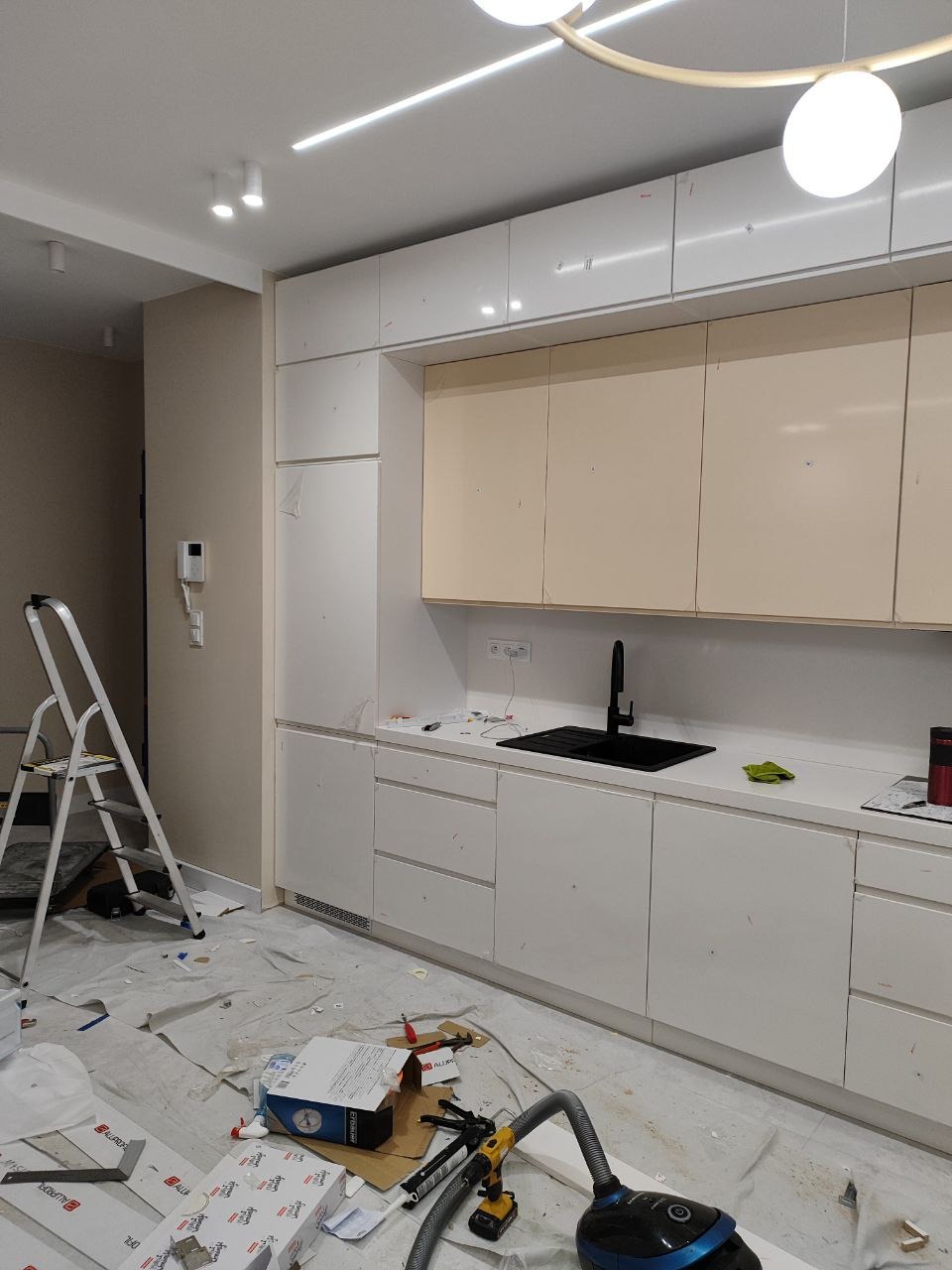 Kuchnia w trakcie montażu: białe szafki górne i dolne, czarny zlew i bateria, zabezpieczona podłoga, drabina i odkurzacz widoczne na pierwszym planie.