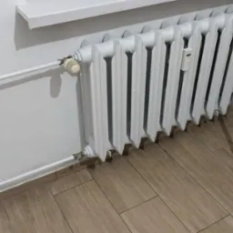 Biały, żeliwny grzejnik członowy z termostatem i elektronicznym miernikiem ciepła, zamontowany na ścianie z drewnianą boazerią i płytkami imitującymi drewno.