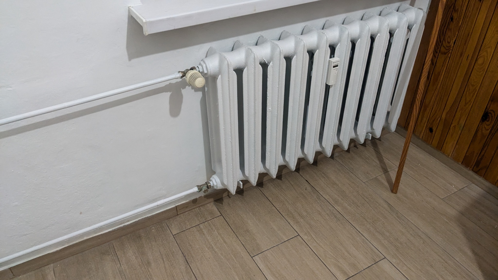 Biały, żeliwny grzejnik członowy z termostatem i elektronicznym miernikiem ciepła, zamontowany na ścianie z drewnianą boazerią i płytkami imitującymi drewno.