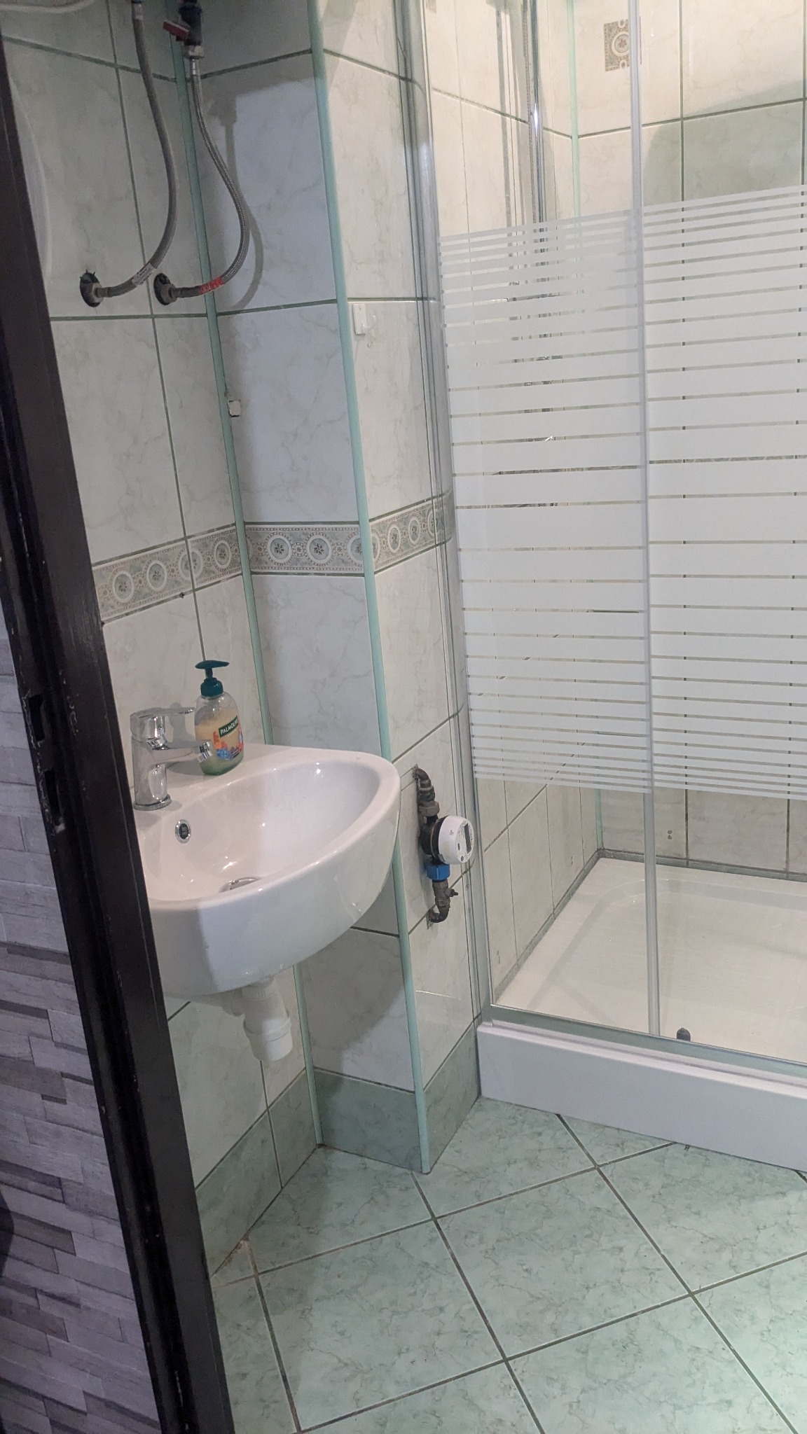 Wykończona łazienka z umywalką, kabiną prysznicową i płytkami w odcieniach zieleni. Widoczne rury doprowadzające wodę i licznik.