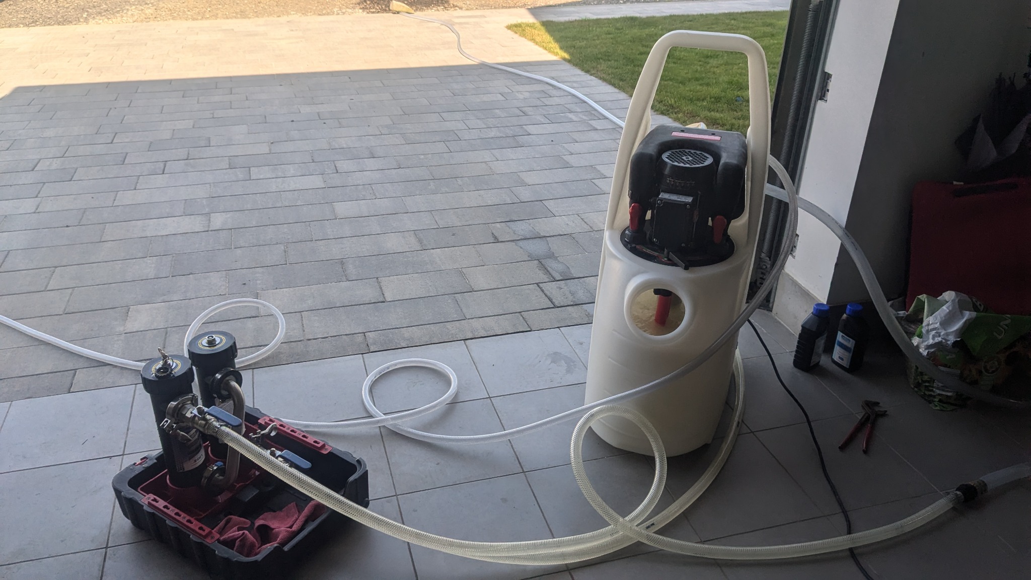 Przenośna pompa do cieczy z filtrami i przezroczystymi wężami na tle garażu z kostki brukowej. Widoczne narzędzia i pojemniki z płynami.