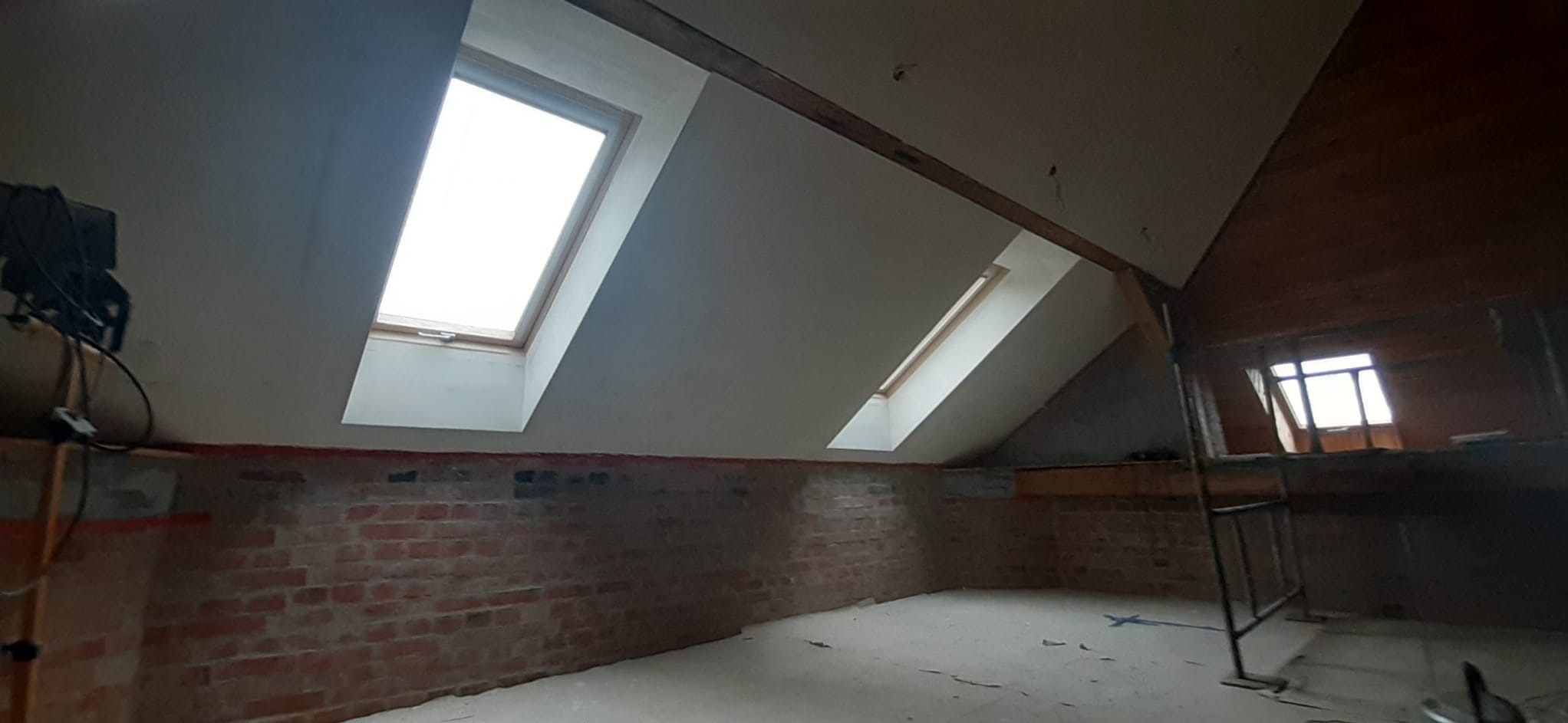Poddasze po remoncie: gładkie, białe ściany z oknami dachowymi, odsłonięta cegła na dole, drewniana konstrukcja dachu. Widać elementy rusztowania.