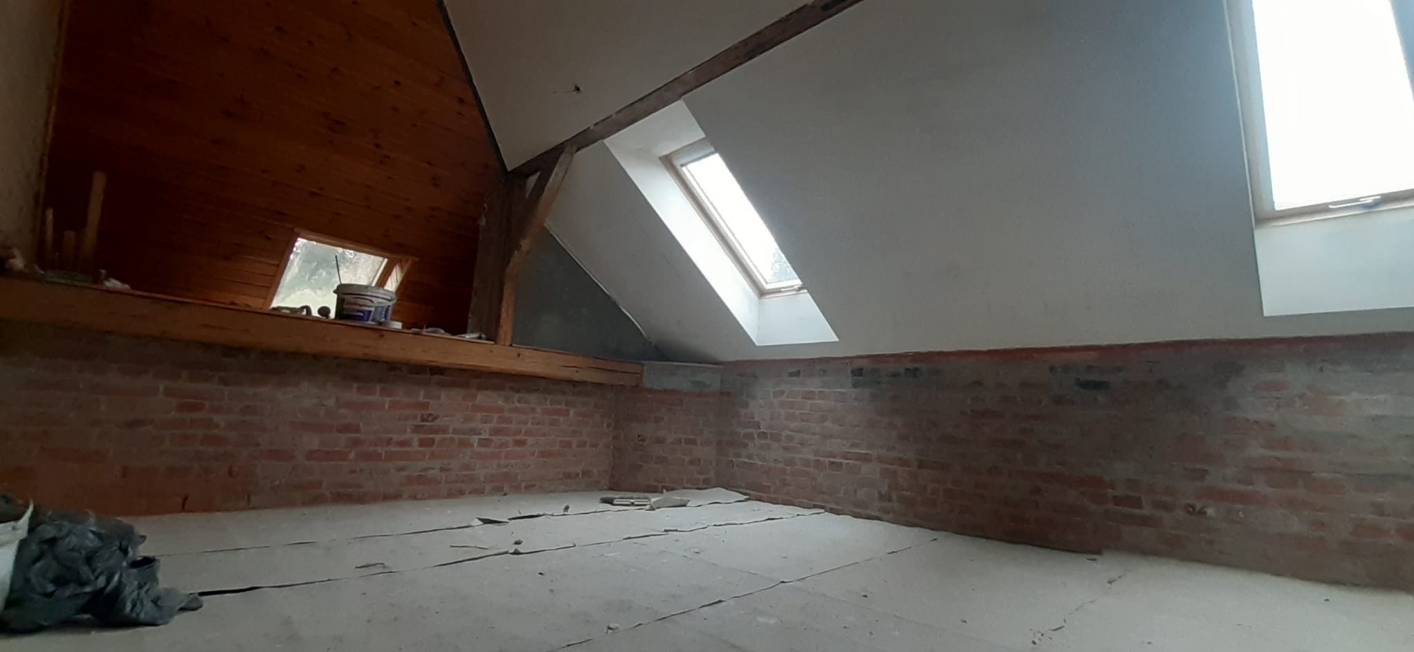 Poddasze z ceglaną ścianą i oknem dachowym po częściowym wykończeniu gładzią. Widoczne drewniane elementy konstrukcyjne i resztki materiałów budowlanych.