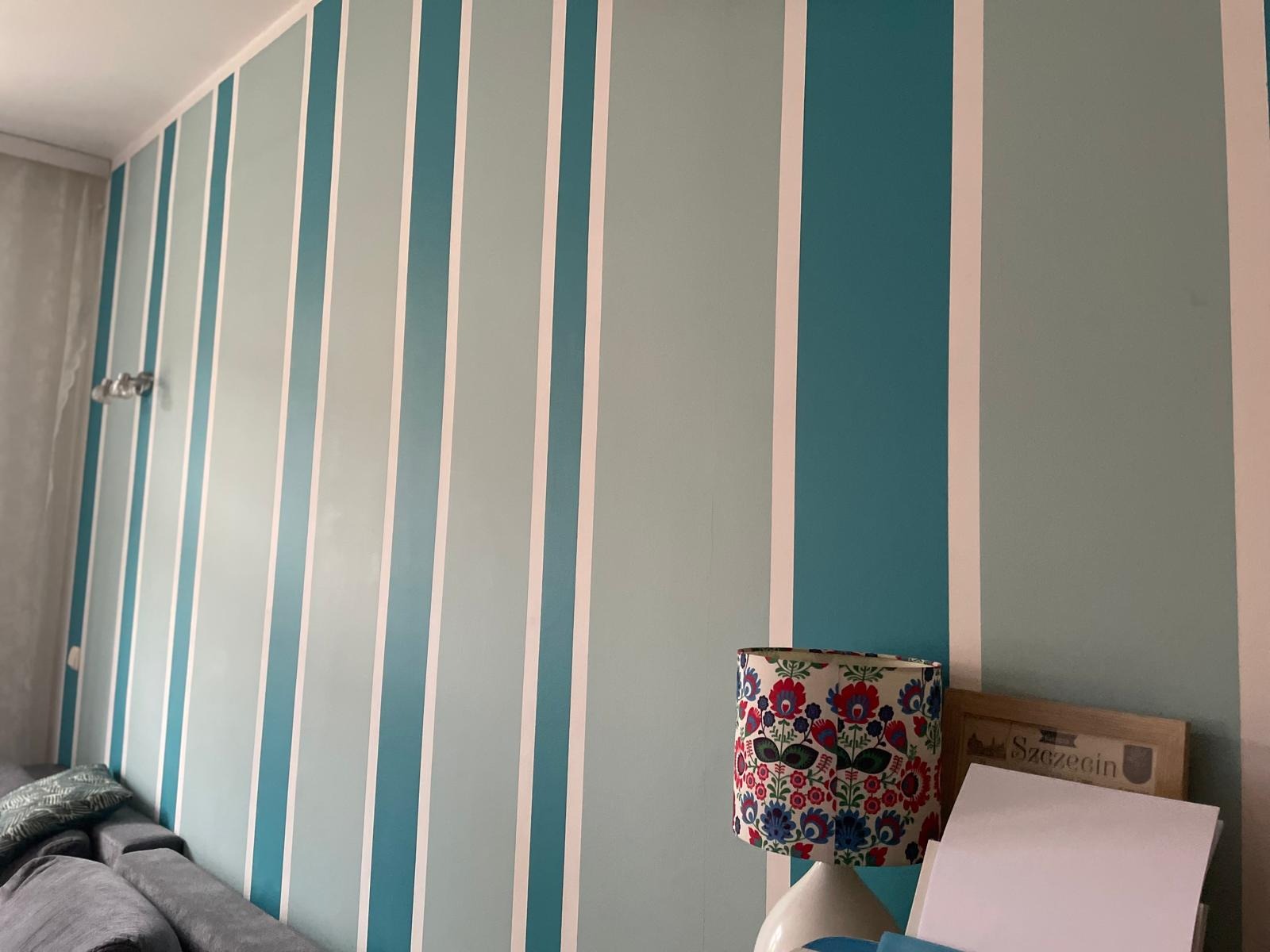 Ściana w paski w odcieniach błękitu i szarości z lampą z folkowym abażurem. Dekoracyjne malowanie ścian w pokoju.