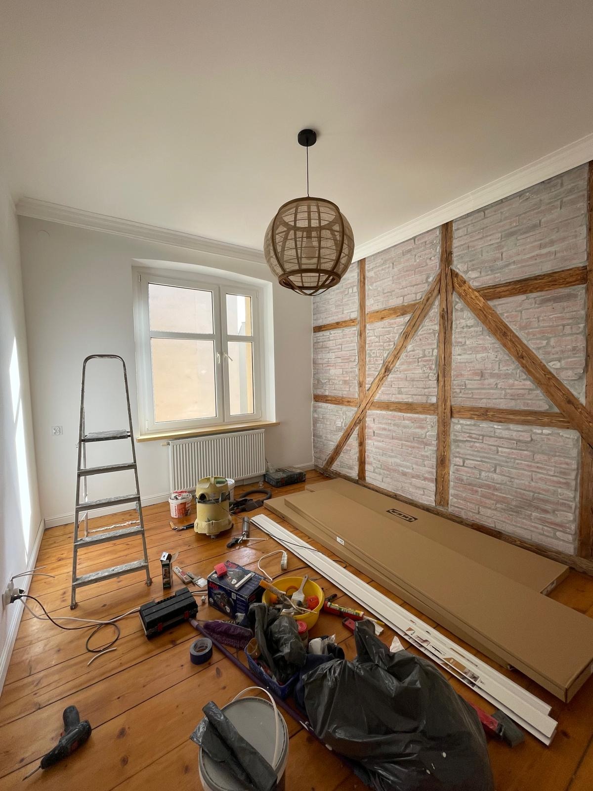 Pokój w trakcie prac wykończeniowych: drabina, narzędzia, panele podłogowe i dekoracyjna ściana z cegły oraz drewnianą konstrukcją. Widoczna lampa wisząca.