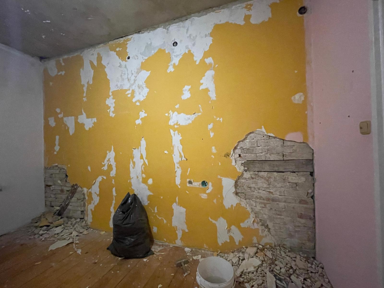 Pokój w trakcie remontu: żółta ściana z resztkami starej farby, odsłonięta cegła, gruz na podłodze, worek na śmieci. Widoczne ślady po gniazdkach elektrycznych.