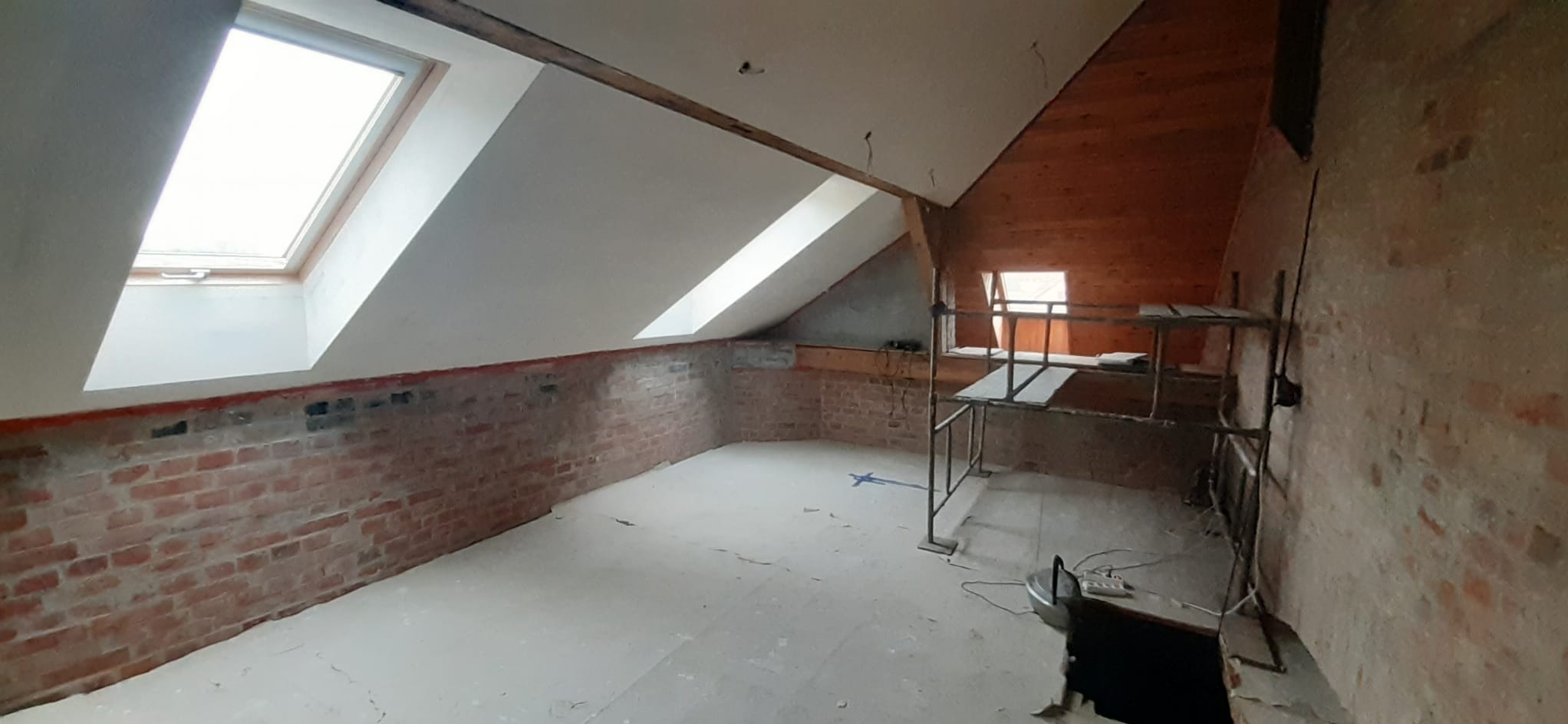 Poddasze w trakcie remontu: surowe cegły, nowe okna dachowe, rusztowanie. Widoczne ślady prac wykończeniowych na ścianach i podłodze, przygotowanie do dalszych etapów.