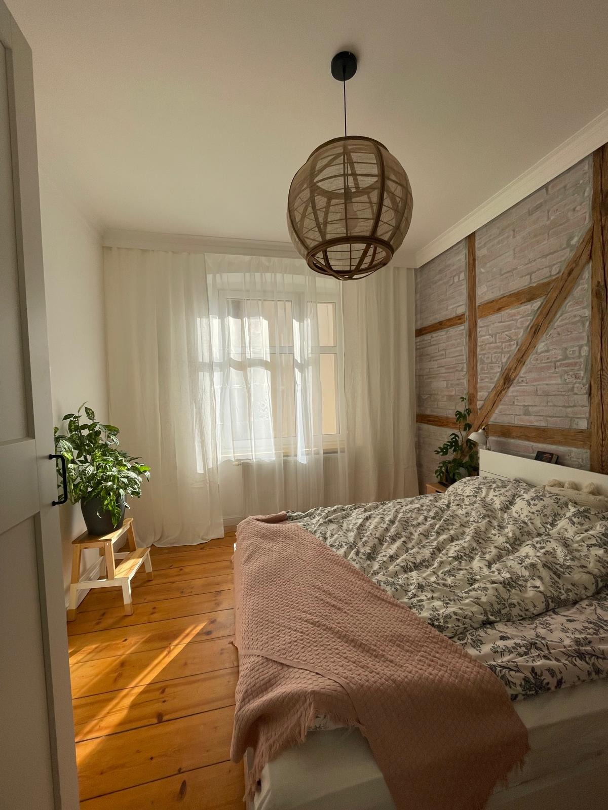 Jasna sypialnia z drewnianą podłogą i ceglaną ścianą. Lampa z wikliny zwisa nad łóżkiem z narzutą w kwiaty. Roślina doniczkowa na drewnianym stołku przy oknie z białymi zasłonami.