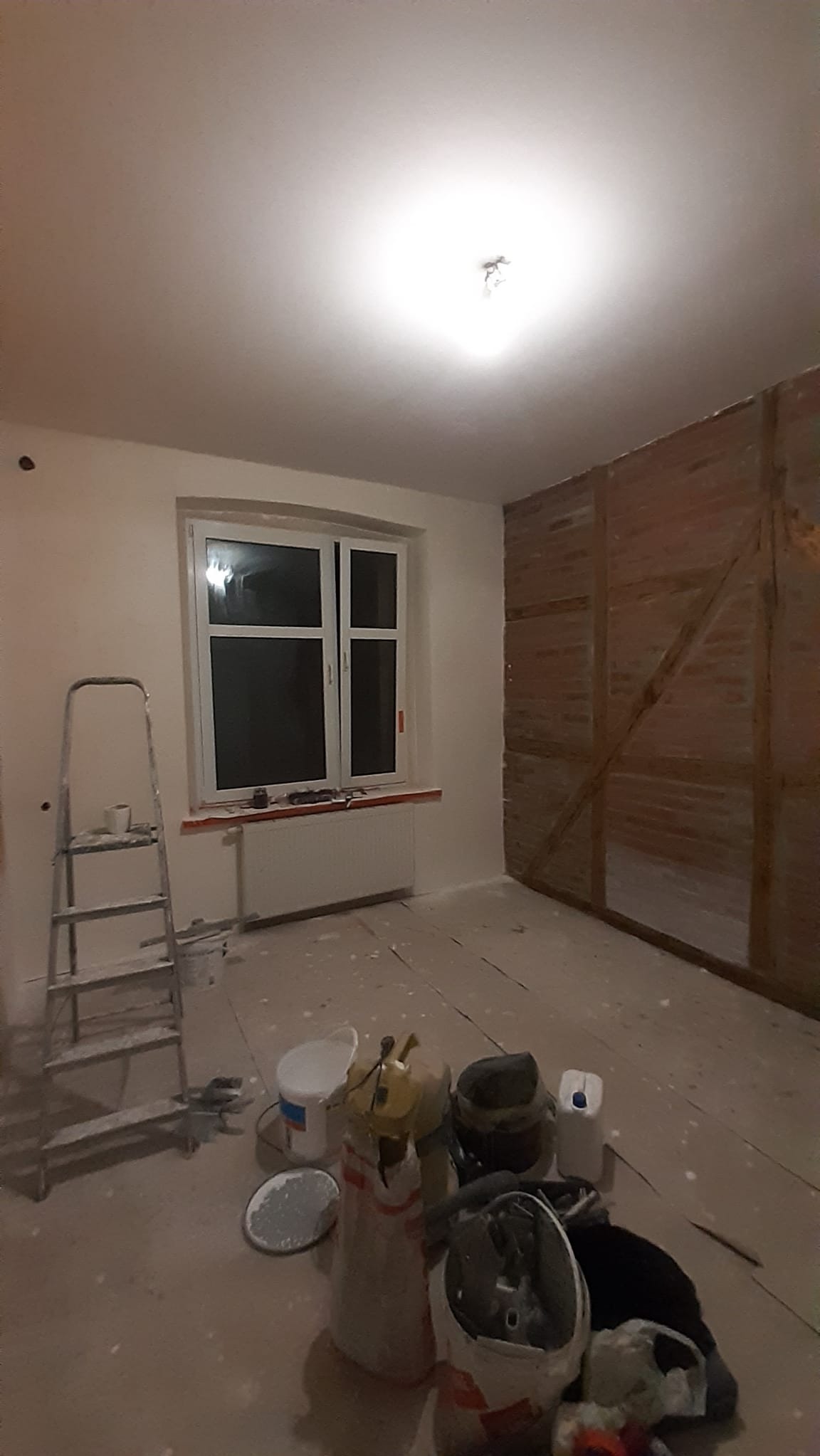 Pokój w trakcie remontu: ściany częściowo pomalowane na biało, drabina, narzędzia malarskie i fragment ściany z cegły. Widoczne ślady farby na podłodze.