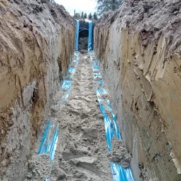 Wykop z ułożonymi niebieskimi rurami wodociągowymi, częściowo zasypany piaskiem, widok z góry na dno wykopu.