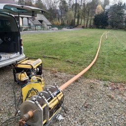 KEEM - Zgrzewanie rur wodociągowych na zewnątrz budynku, widoczna zgrzewarka, agregat prądotwórczy, otwarta paka samochodu dostawczego i ułożony odcinek rury na trawniku.