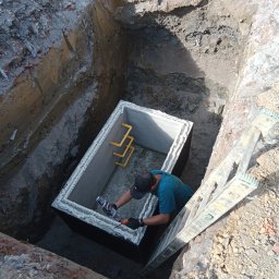 KEEM - Pracownik w wykopie uszczelnia betonowy zbiornik szarą pianką montażową, widoczna drabina oparta o krawędź wykopu.