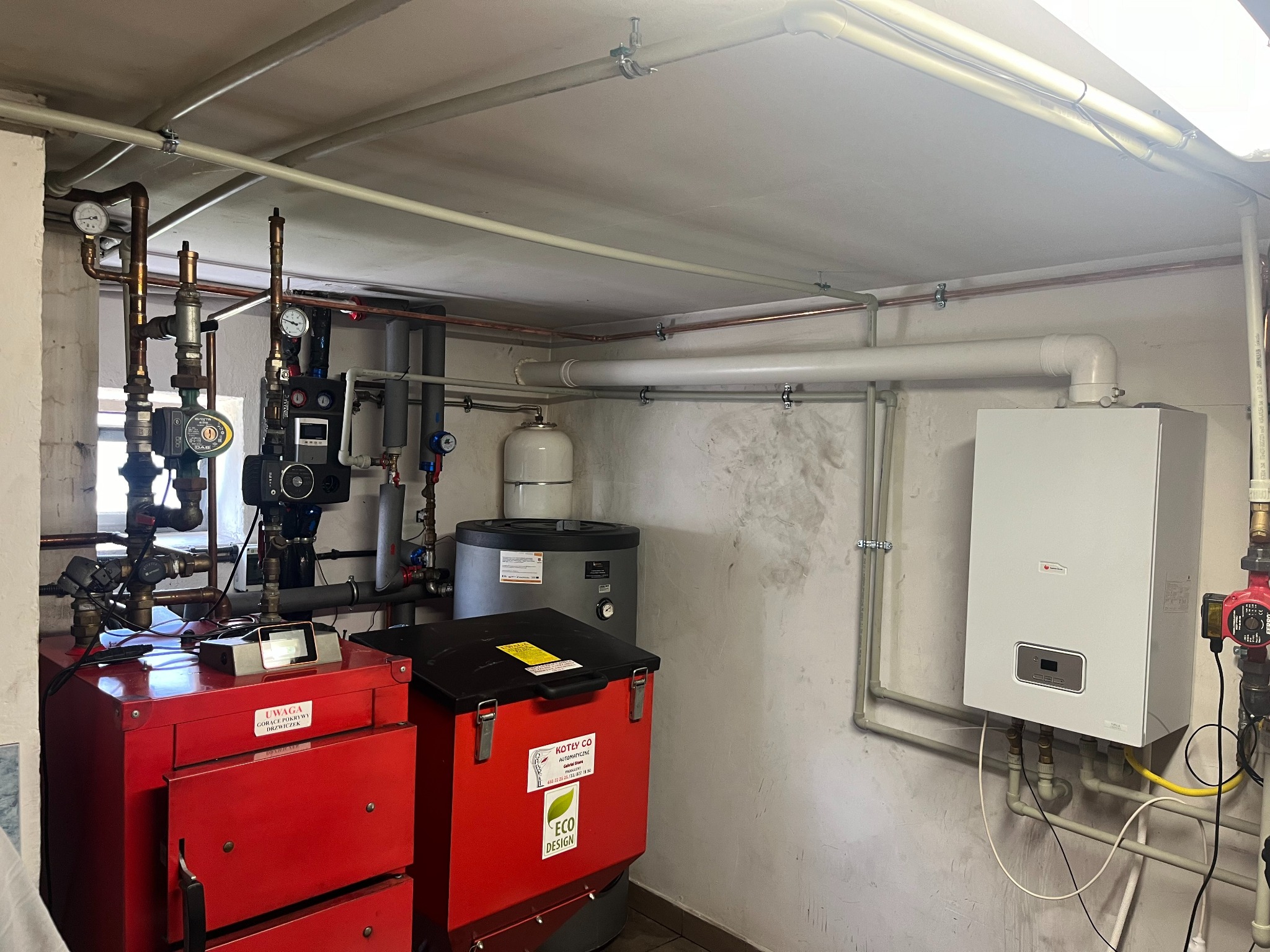 Kotłownia z czerwonym kotłem na ekogroszek z napisem ECO DESIGN, białym piecem gazowym, zbiornikiem buforowym i instalacją grzewczą z rur miedzianych i plastikowych.