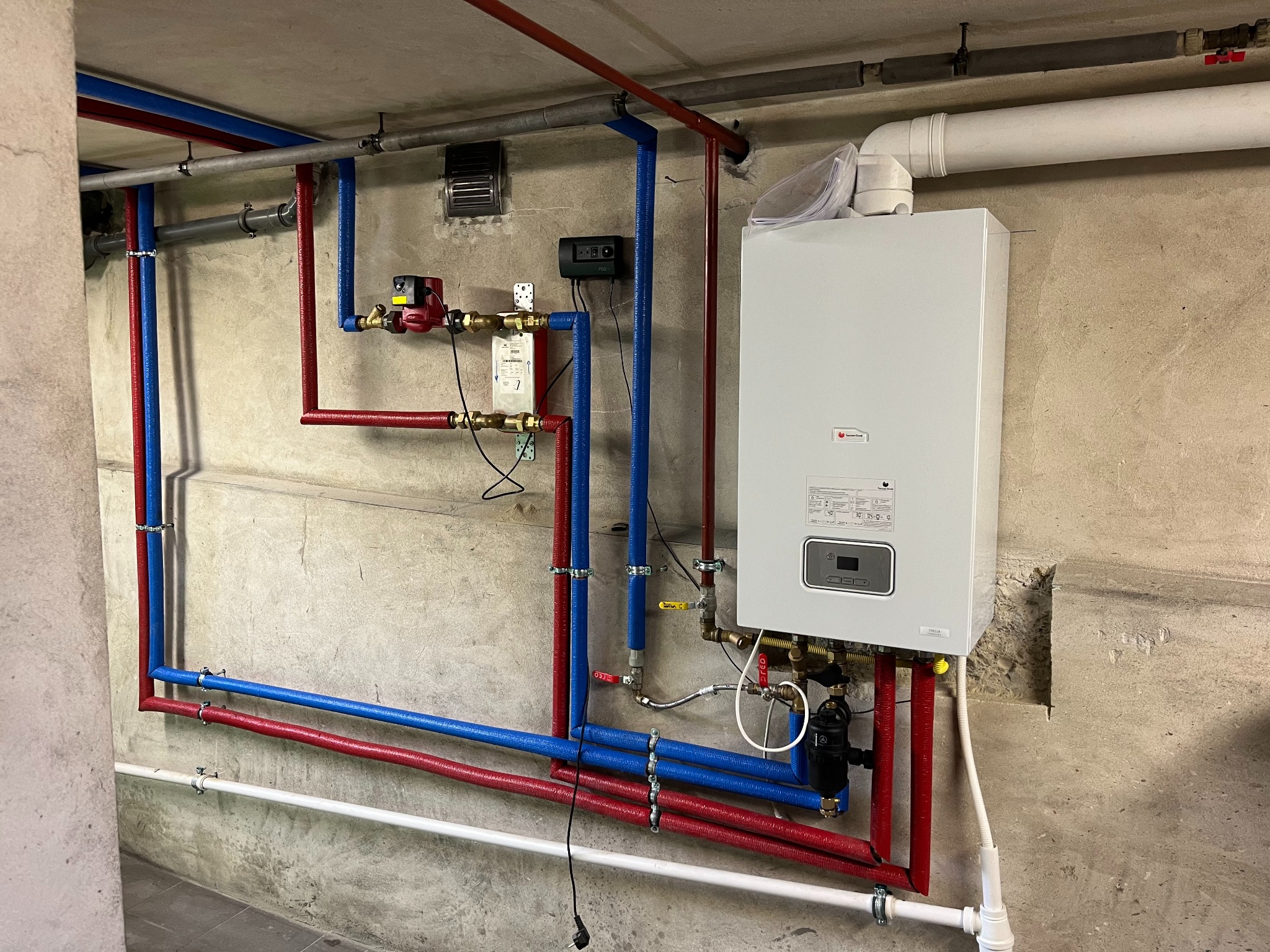 Nowoczesny piec gazowy marki Saunier Duval zamontowany na surowej, betonowej ścianie piwnicy, z widocznymi rurami instalacji grzewczej w kolorach czerwonym i niebieskim, zabezpieczonymi izolacją...