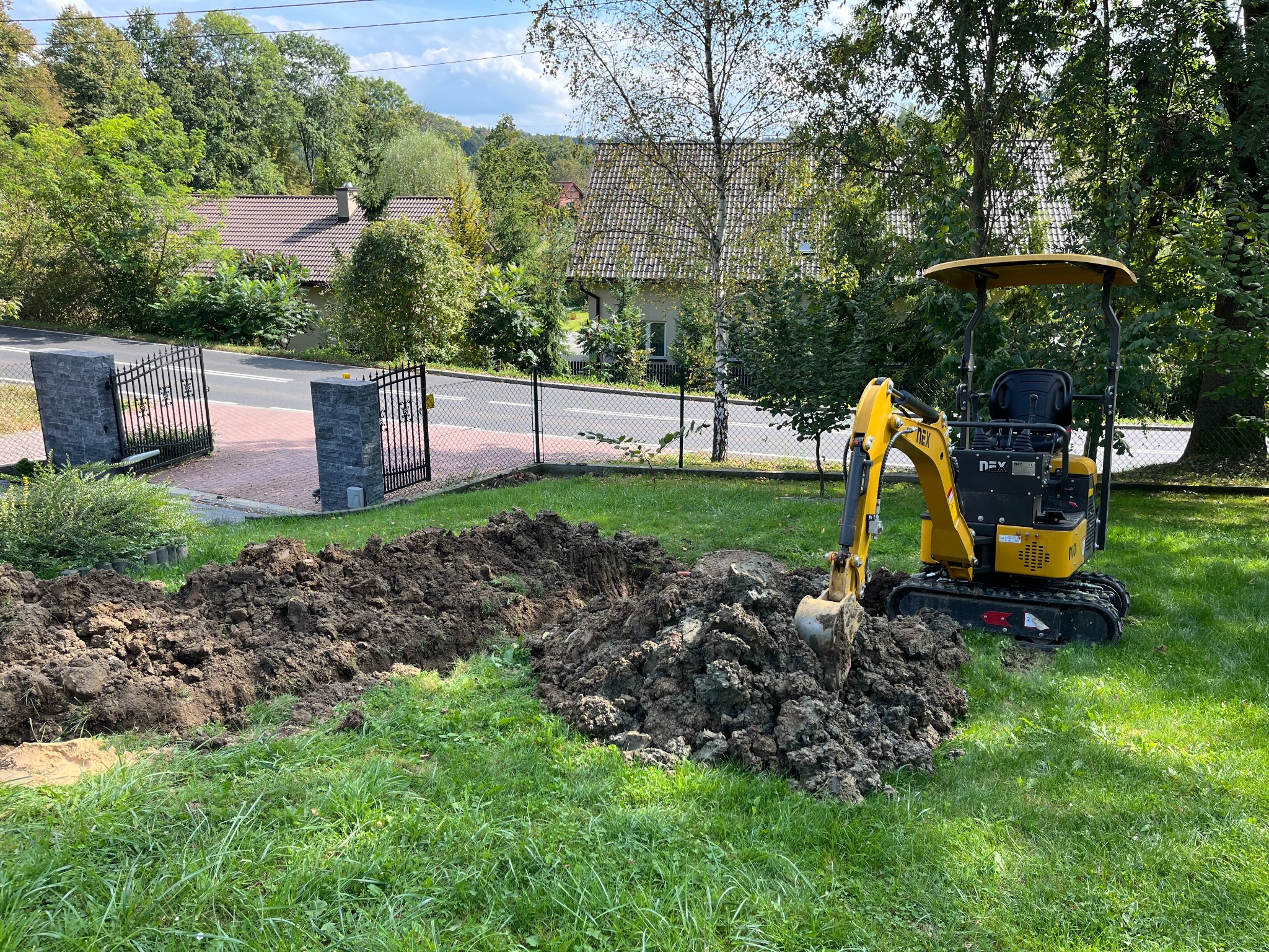 Żółta mini-koparka na trawniku obok wykopanego rowu, w tle ogrodzenie i domy jednorodzinne.