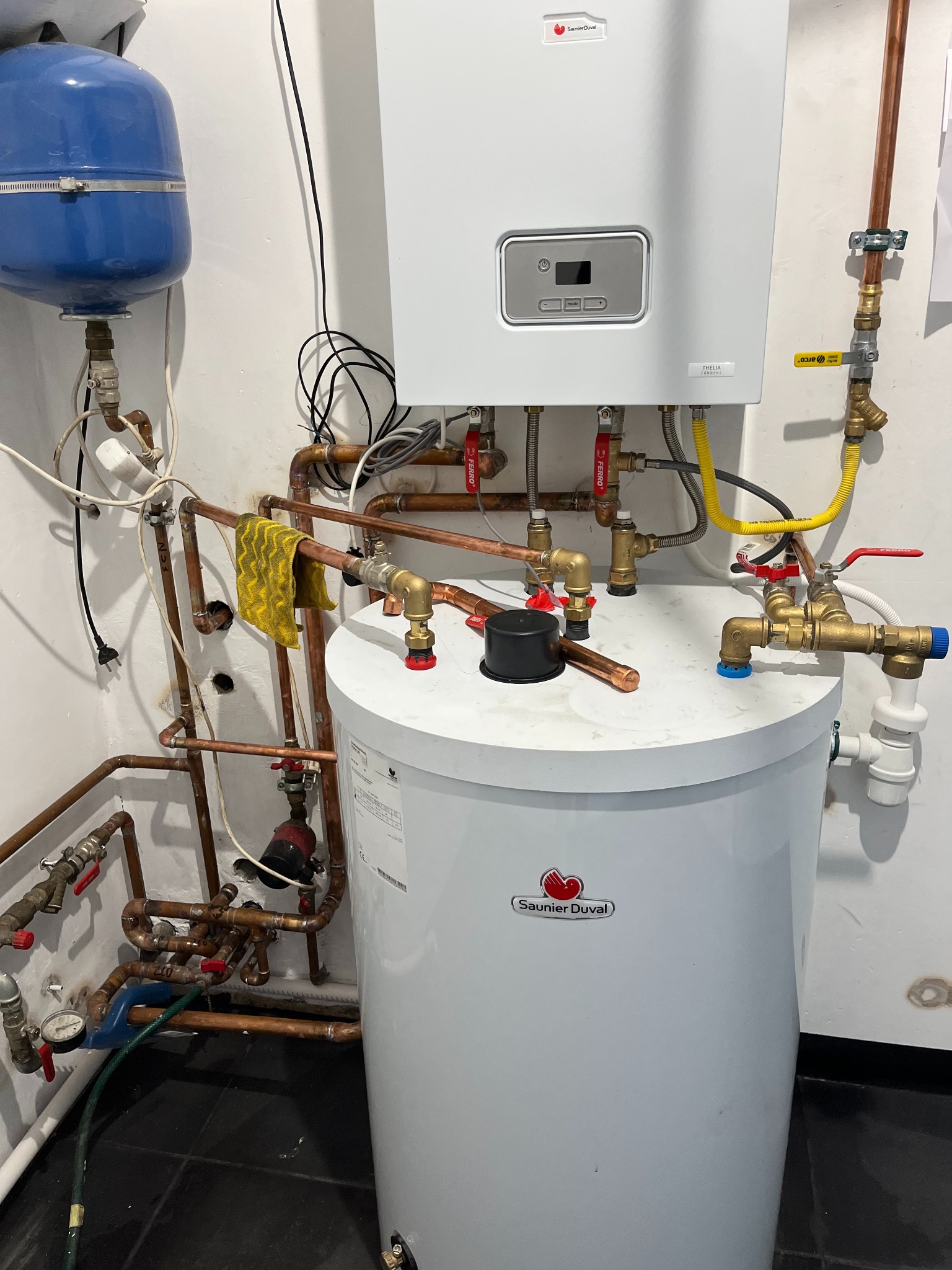 Biały piec gazowy Saunier Duval z licznymi miedzianymi rurami, zaworami i osprzętem w kotłowni; żółta szmatka wisząca na jednej z rur.