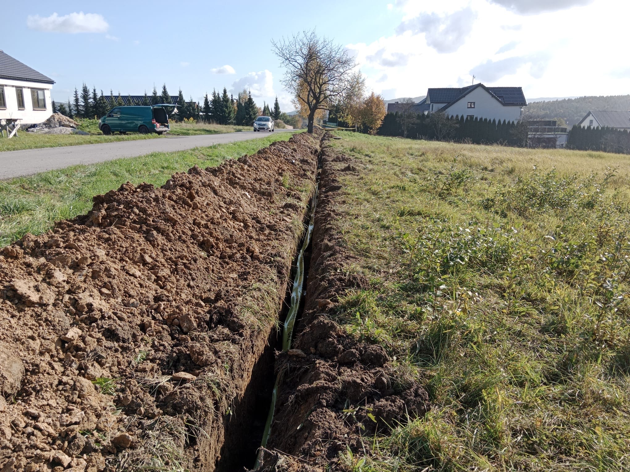 Wykopany rów z widocznymi rurami instalacji podziemnej na tle wiejskiej zabudowy i zielonego busa.
