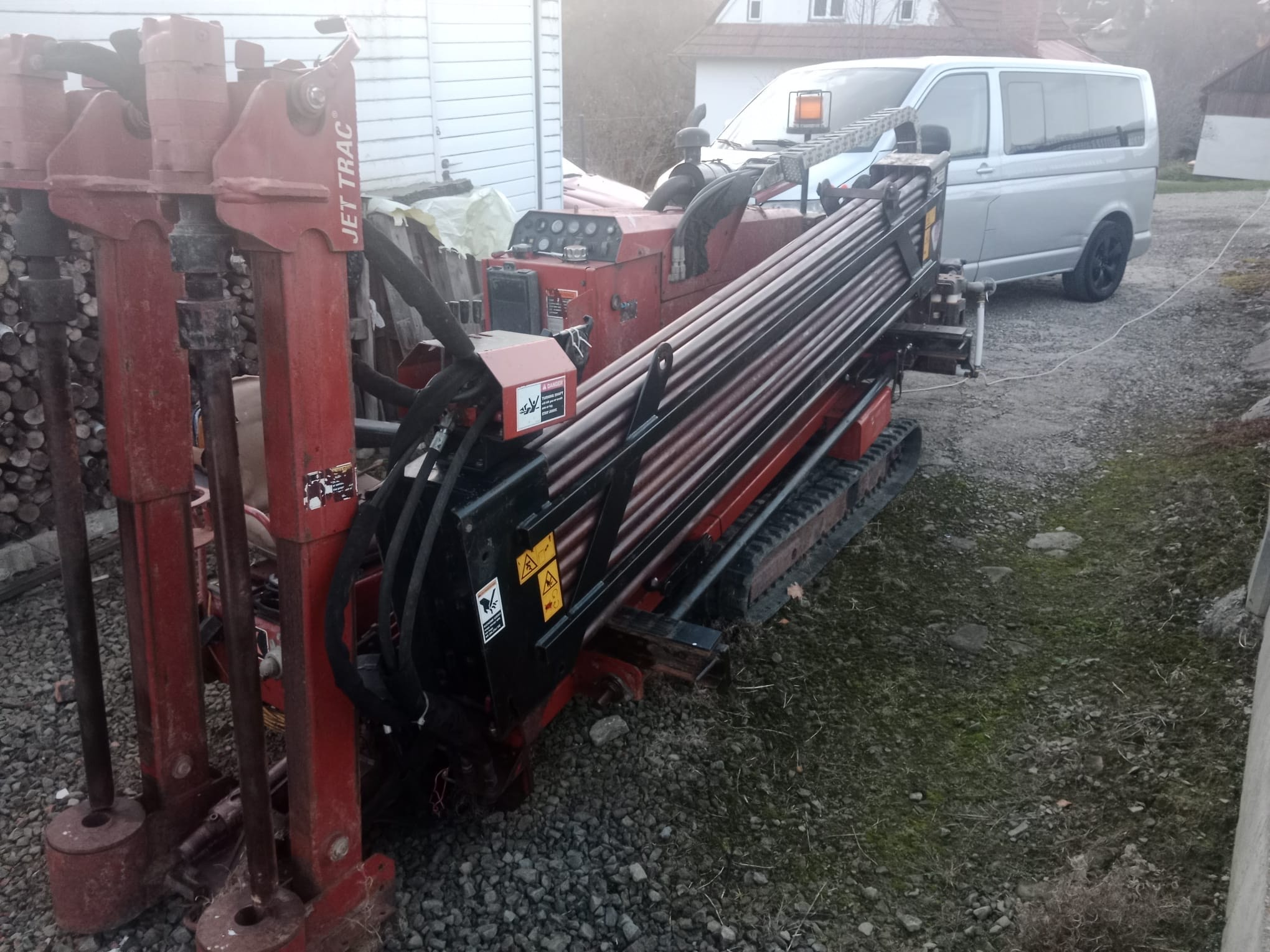 Czerwona wiertnica Jet Trac na gąsienicach z zestawem rur w otoczeniu żwiru i trawy, zaparkowana obok srebrnego vana.