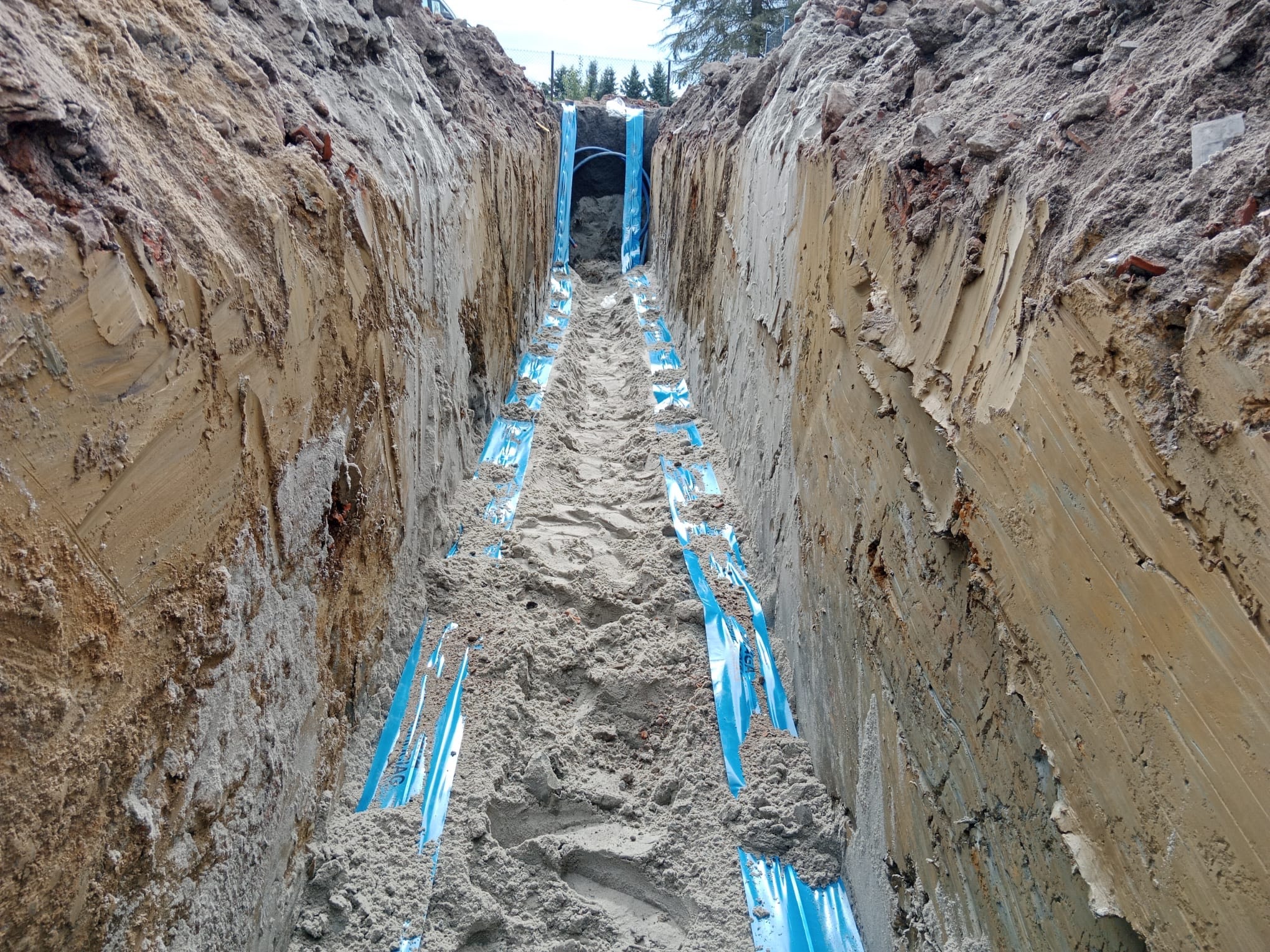 Wykop z ułożonymi niebieskimi rurami wodociągowymi, częściowo zasypany piaskiem, widok z góry na dno wykopu.