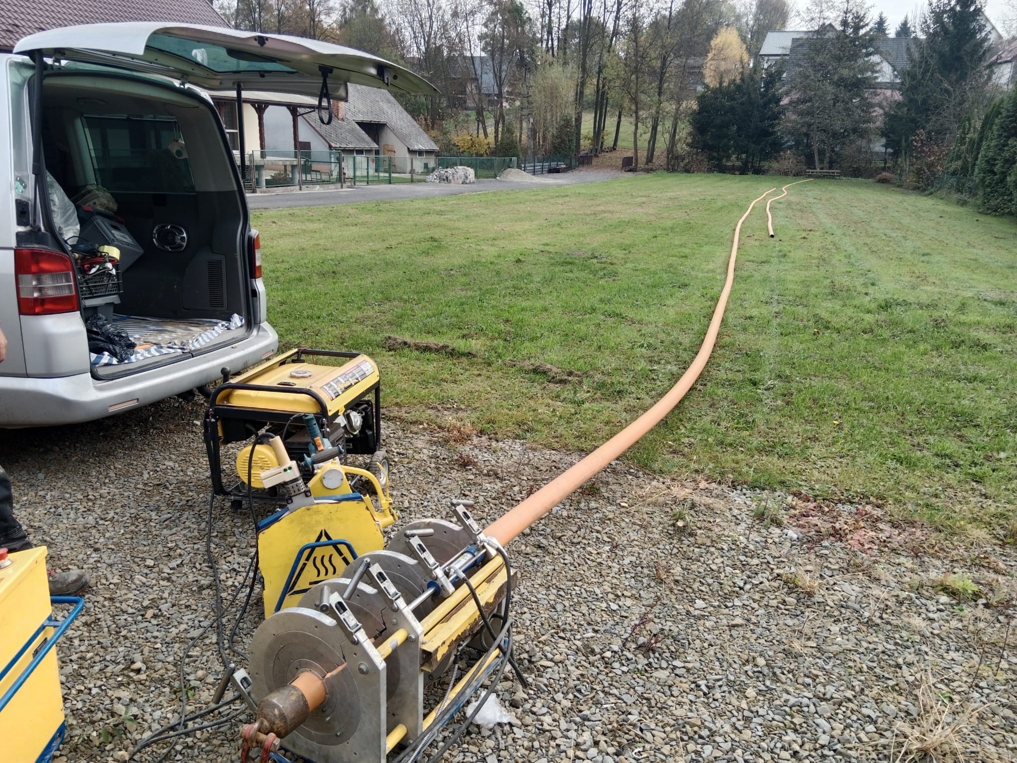 Zgrzewanie rur wodociągowych na zewnątrz budynku, widoczna zgrzewarka, agregat prądotwórczy, otwarta paka samochodu dostawczego i ułożony odcinek rury na trawniku.