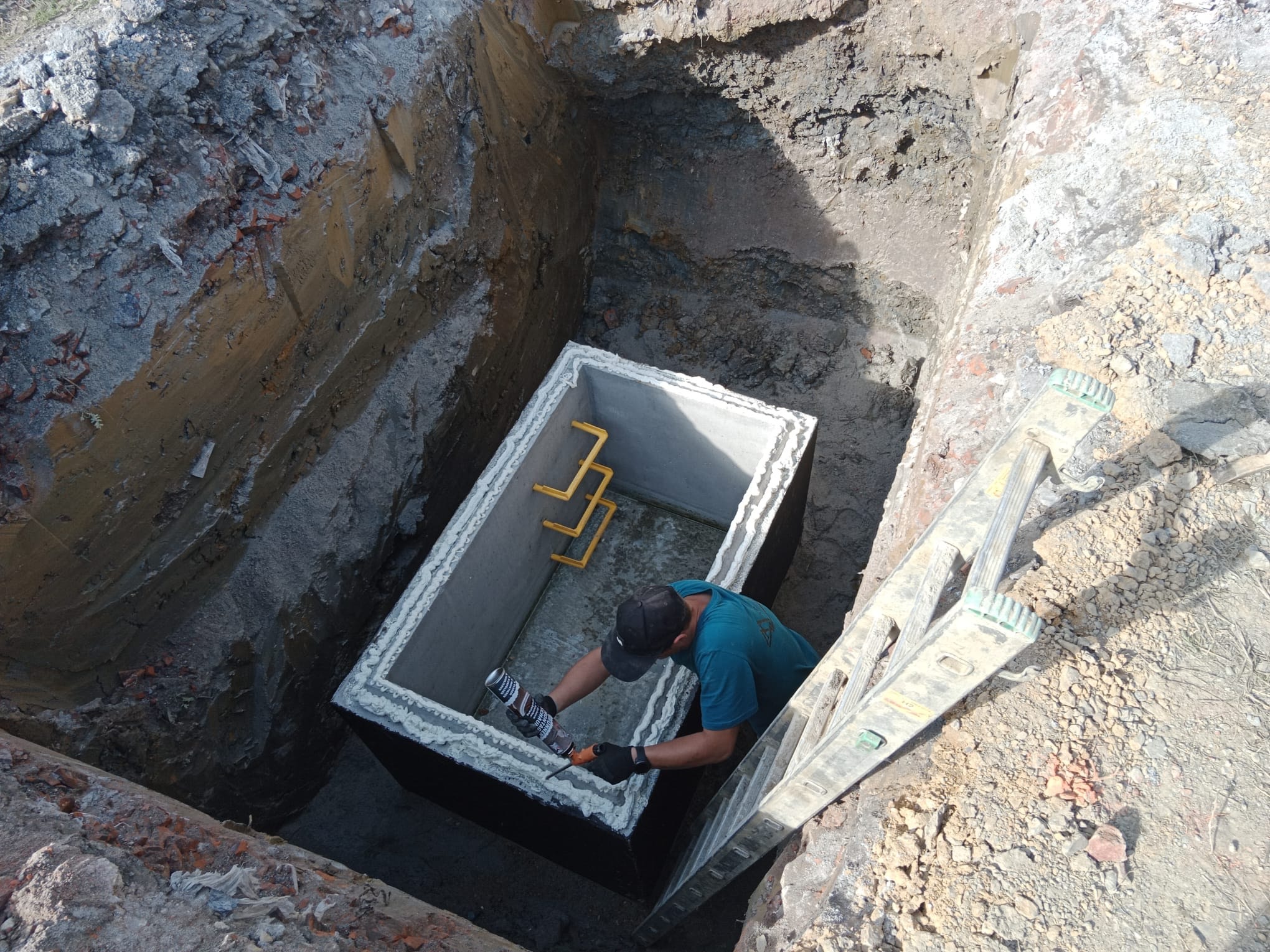 Pracownik w wykopie uszczelnia betonowy zbiornik szarą pianką montażową, widoczna drabina oparta o krawędź wykopu.