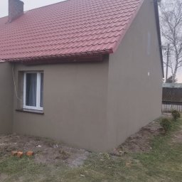 Firma Ogólnobudowlana - Elewacja domu jednorodzinnego po nałożeniu gładzi, widoczna czerwona blachodachówka i białe okno z firanką.