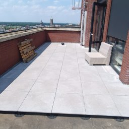Firma Ogólnobudowlana - Wyłożony jasnoszarymi płytami taras na dachu budynku z widokiem na miasto, widoczne elementy regulujące wysokość płyt, paleta oparta o ceglaną ścianę, krzesło z rattanu w rogu.