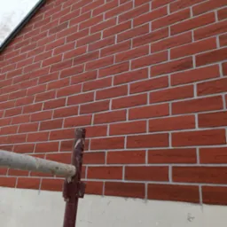 Fragment ściany zewnętrznej budynku z cegły klinkierowej, widoczne rusztowanie malowane na brązowo, podłoże pod cegłą szare.
