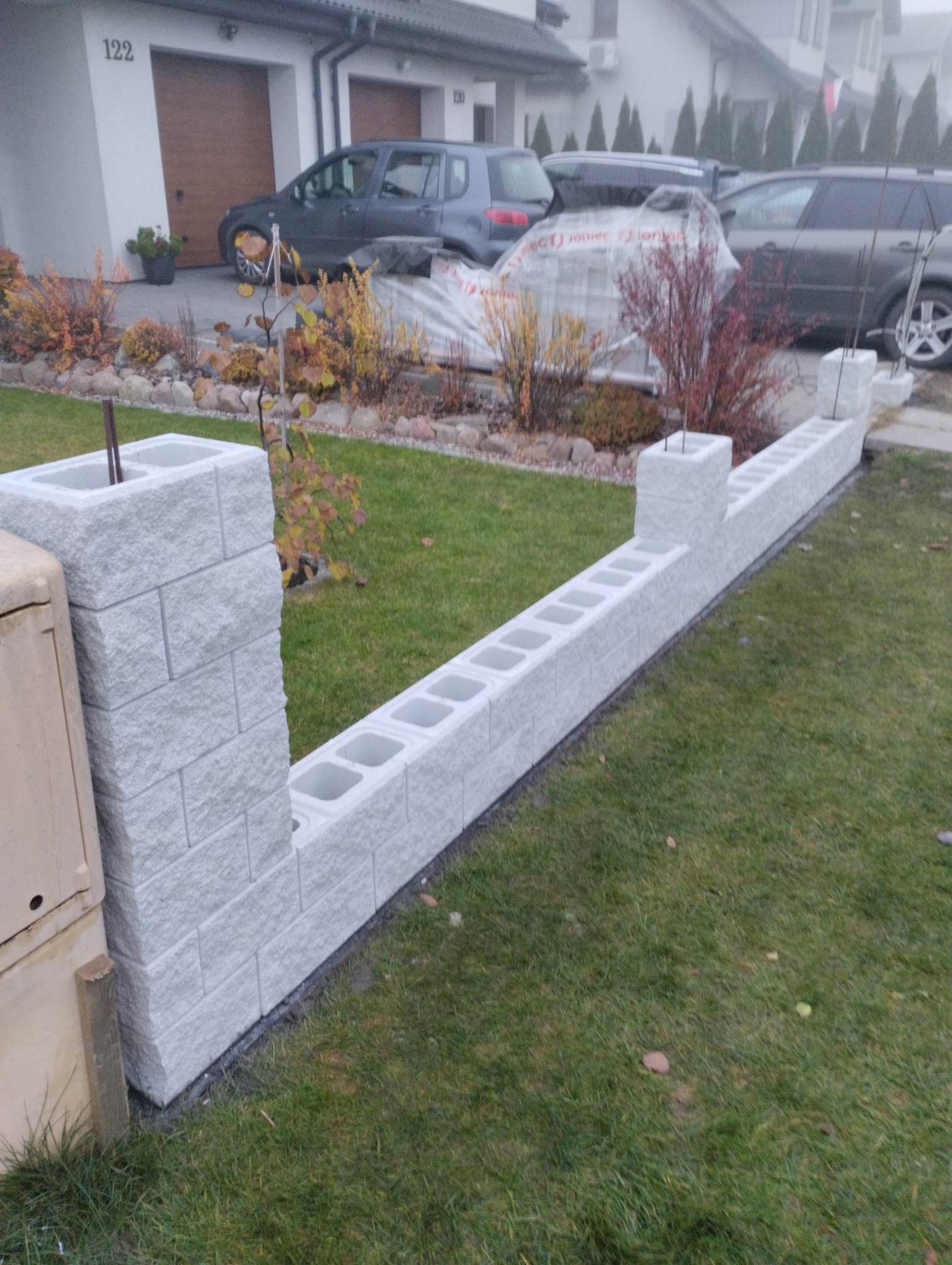 Wznoszenie niskiego muru z szarych betonowych bloczków na trawniku przed domem jednorodzinnym, widoczne zbrojenie w słupkach.