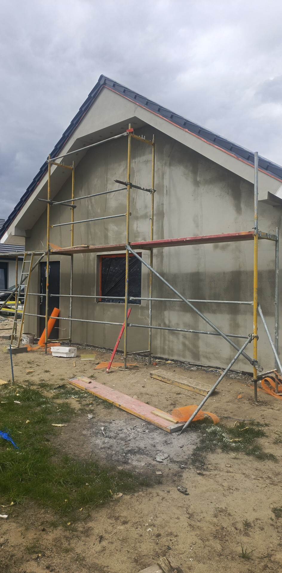 Świeżo otynkowany dom jednorodzinny w trakcie prac wykończeniowych, widoczne rusztowanie, okno zabezpieczone folią, elementy ocieplenia i narzędzia budowlane na ziemi.