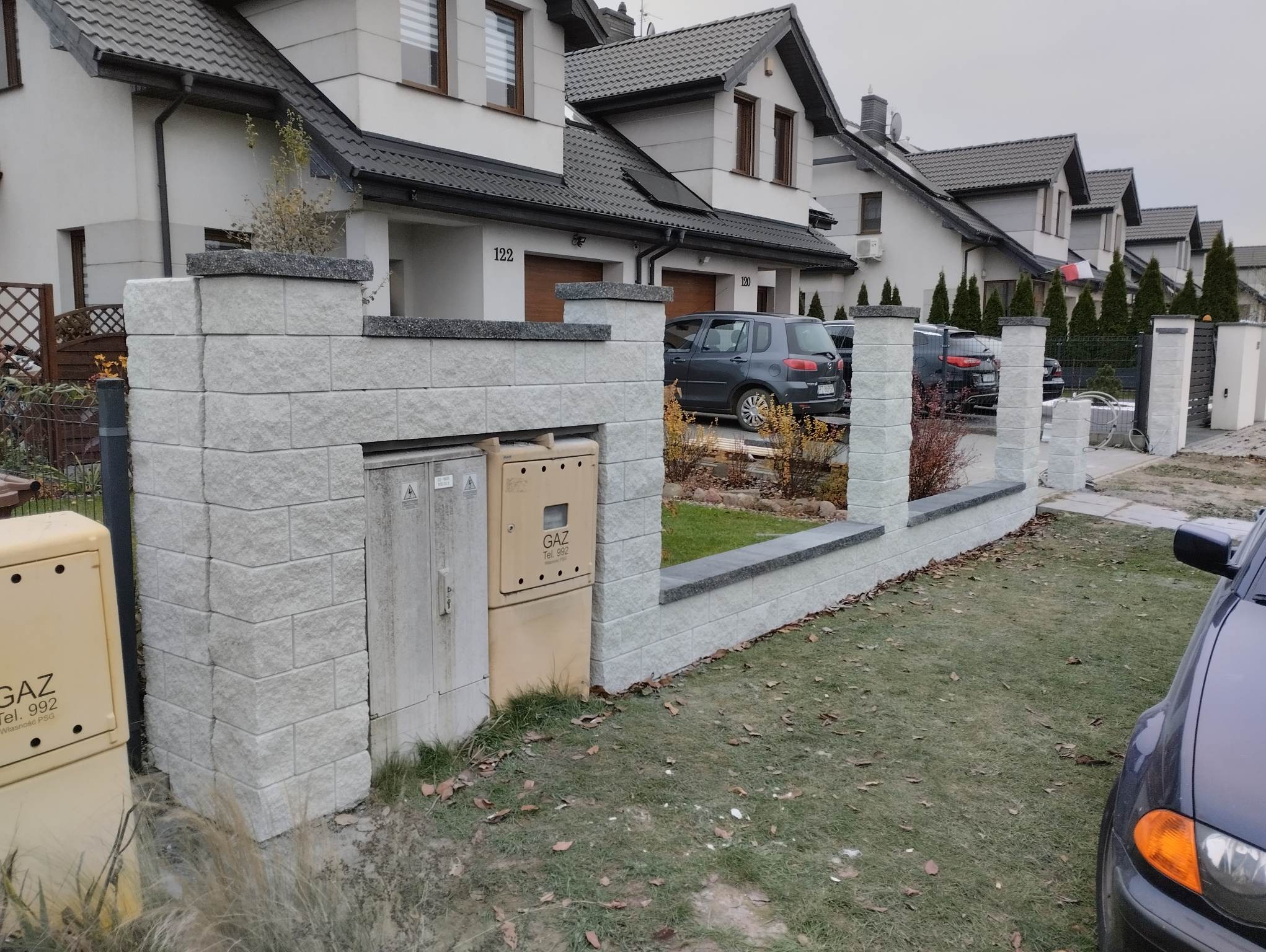 Nowoczesne ogrodzenie z jasnych, blokowych elementów betonowych z ciemnymi nakrywami, otaczające osiedle domów jednorodzinnych z szarą elewacją i ciemnymi dachami, skrzynka gazowa przy ogrodzeniu.