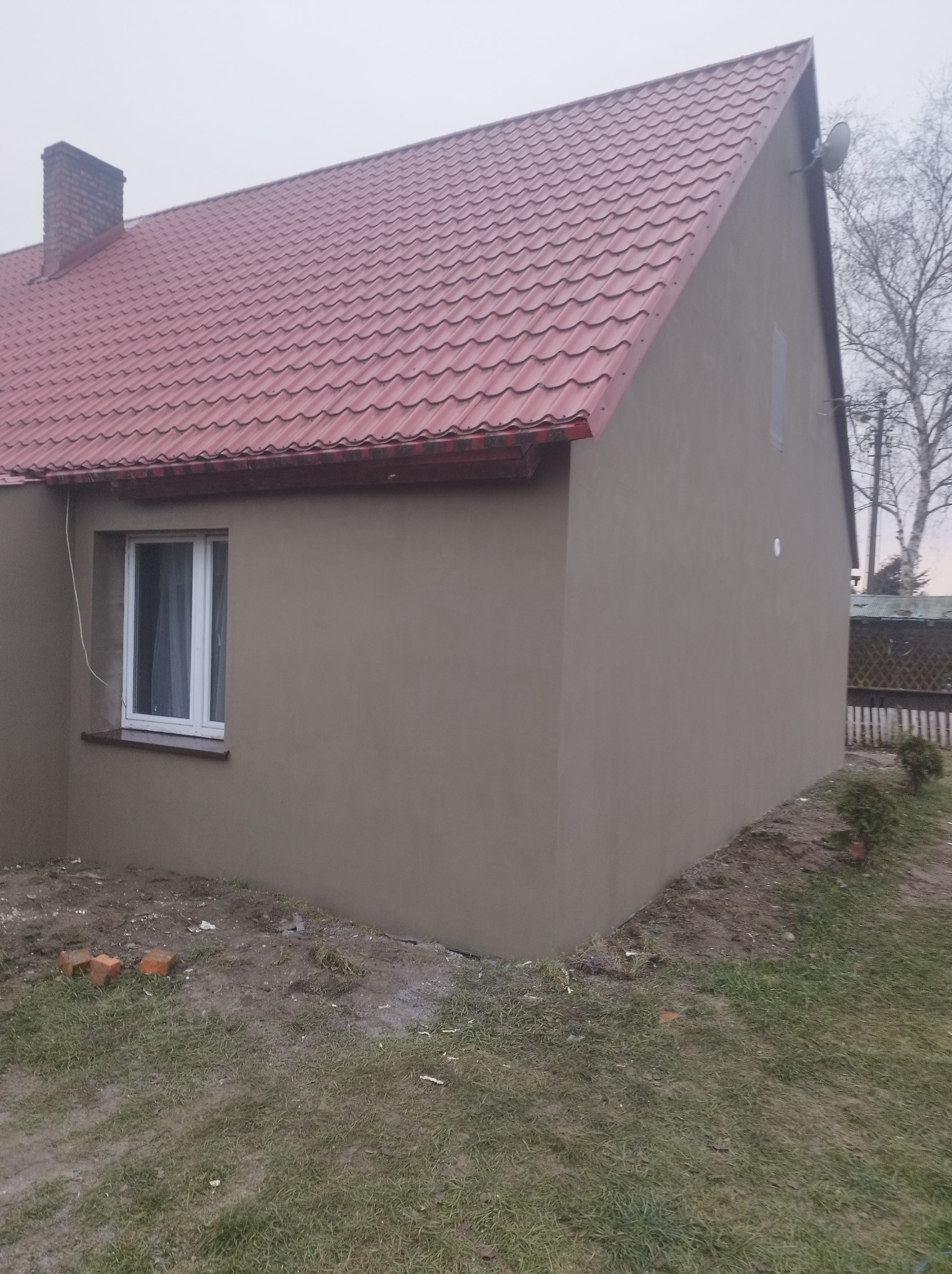 Elewacja domu jednorodzinnego po nałożeniu gładzi, widoczna czerwona blachodachówka i białe okno z firanką.