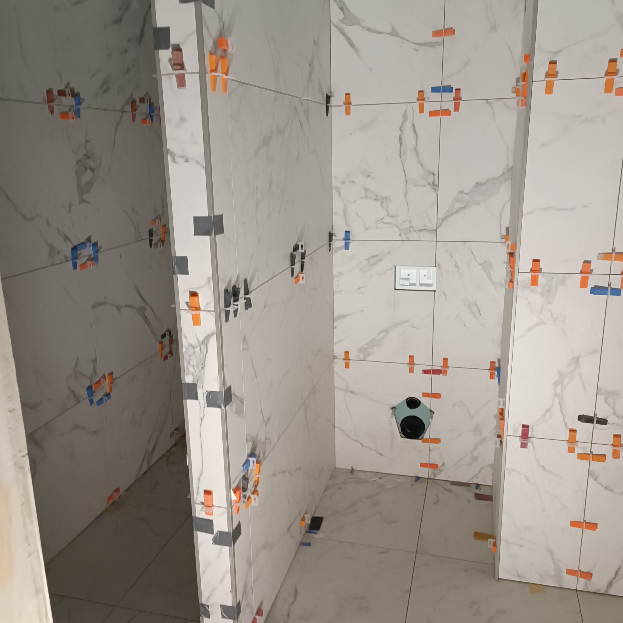 Białe marmurowe płytki ścienne i podłogowe w trakcie montażu, z użyciem klipsów poziomujących i sznurka, widoczny otwór na odpływ.