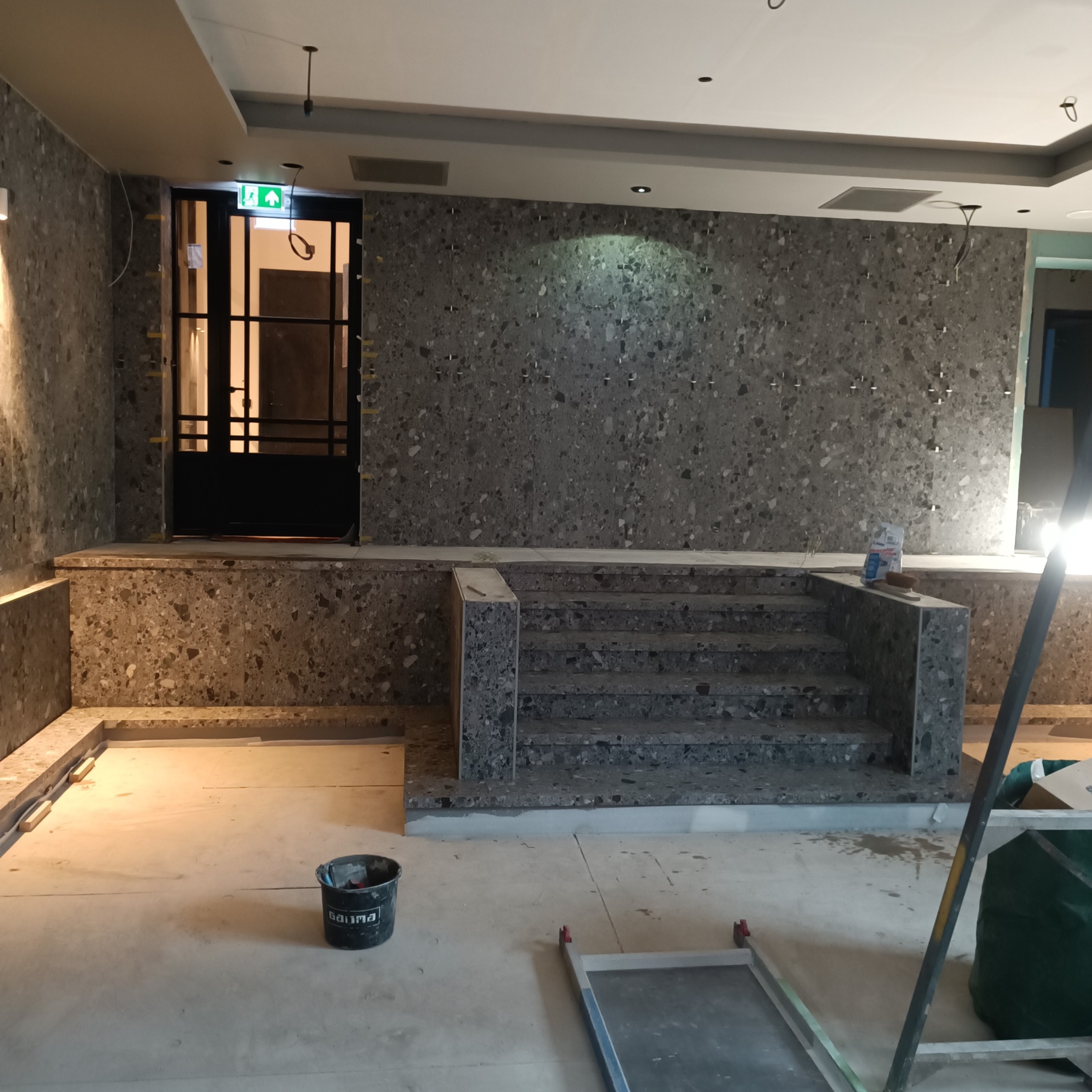 Montaż schodów i okładziny ściennej z płytek lastryko w przestrzeni komercyjnej, widoczny czarny kubeł, narzędzia i zaplecze budowlane.