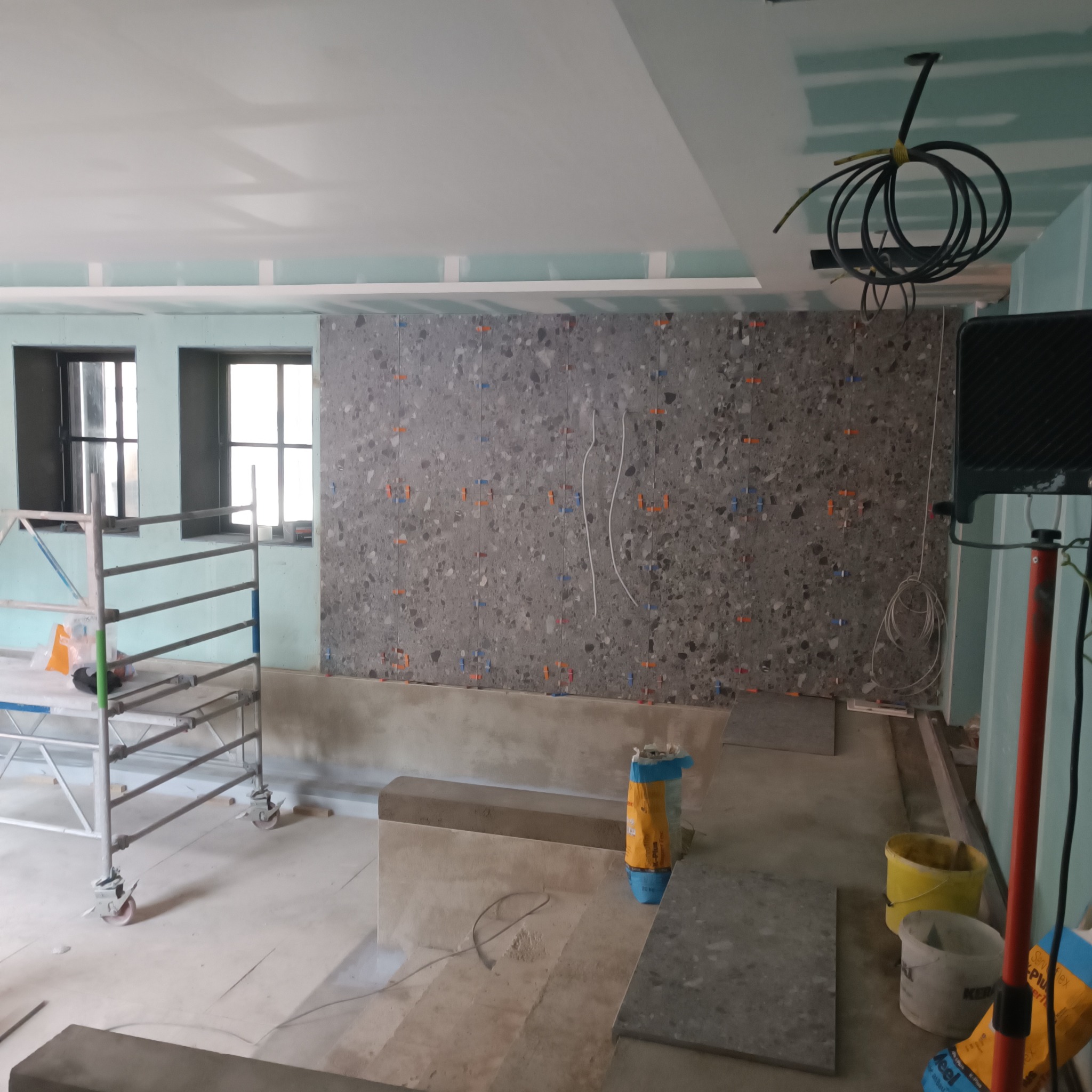 Wnętrze w trakcie prac wykończeniowych, ściana z szarych płytek z kolorowymi drobinkami, krzyżyki dystansowe, widoczne przewody elektryczne, rusztowanie i narzędzia.