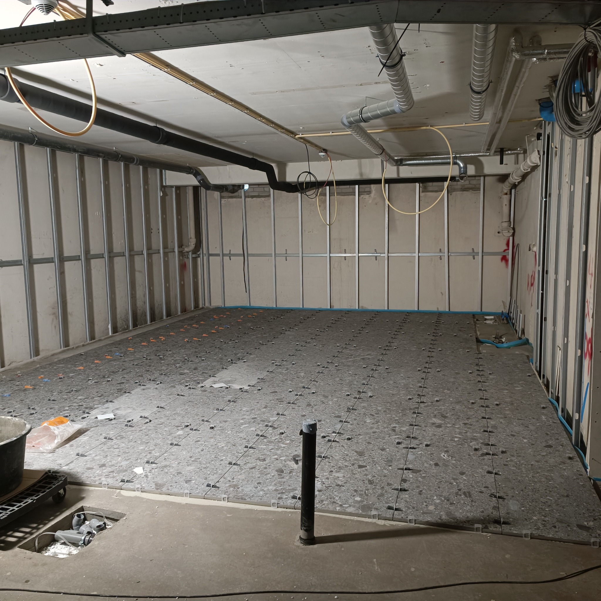 Układanie szarej terazzo podłogi w pomieszczeniu ze stelażem ścian działowych, widoczne rury i elementy instalacji pod sufitem.