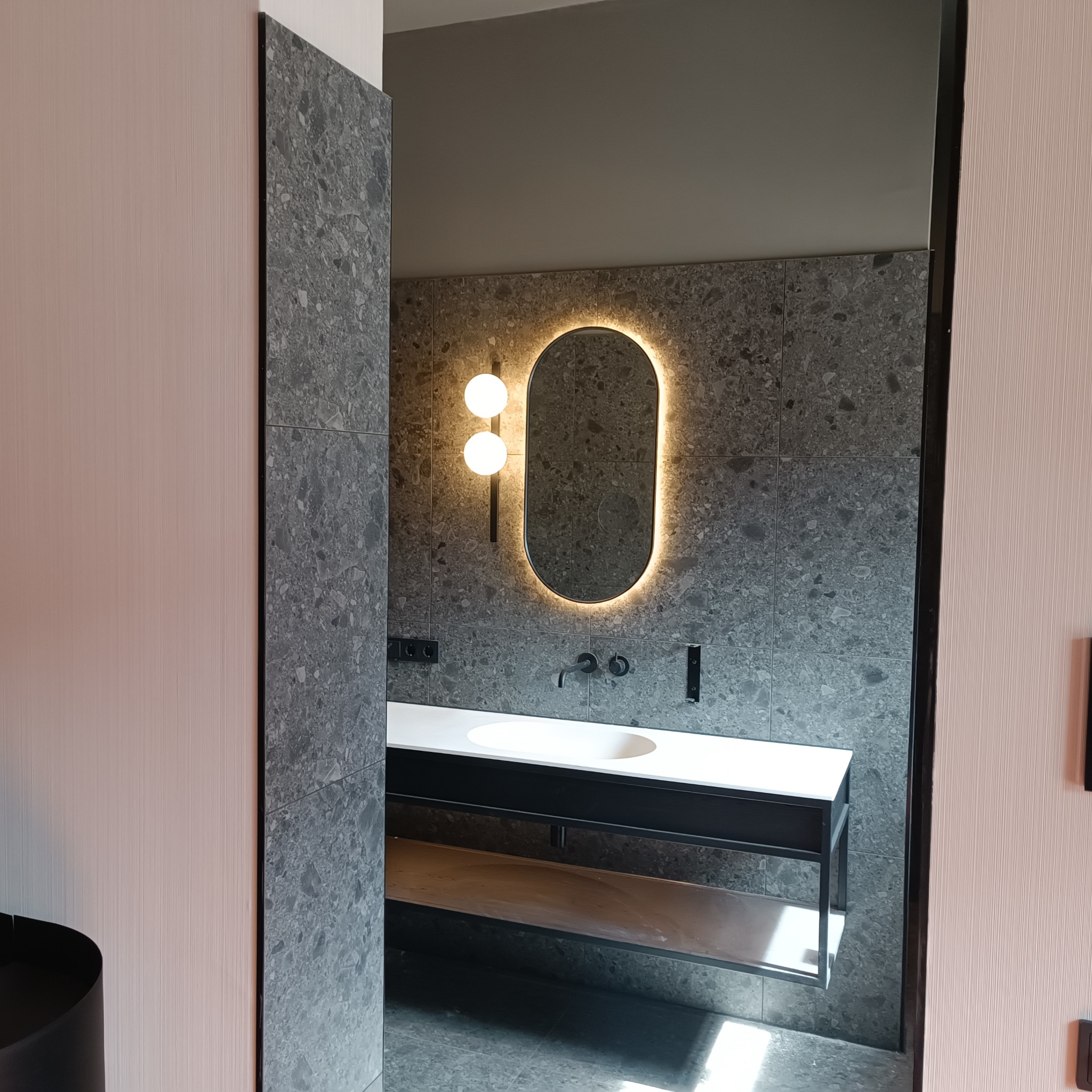 Nowoczesna łazienka z szarymi płytkami lastryko, owalnym lustrem z podświetleniem LED, białą umywalką na czarnym stelażu i minimalistyczną armaturą, widziana zza częściowo otwartych drzwi.