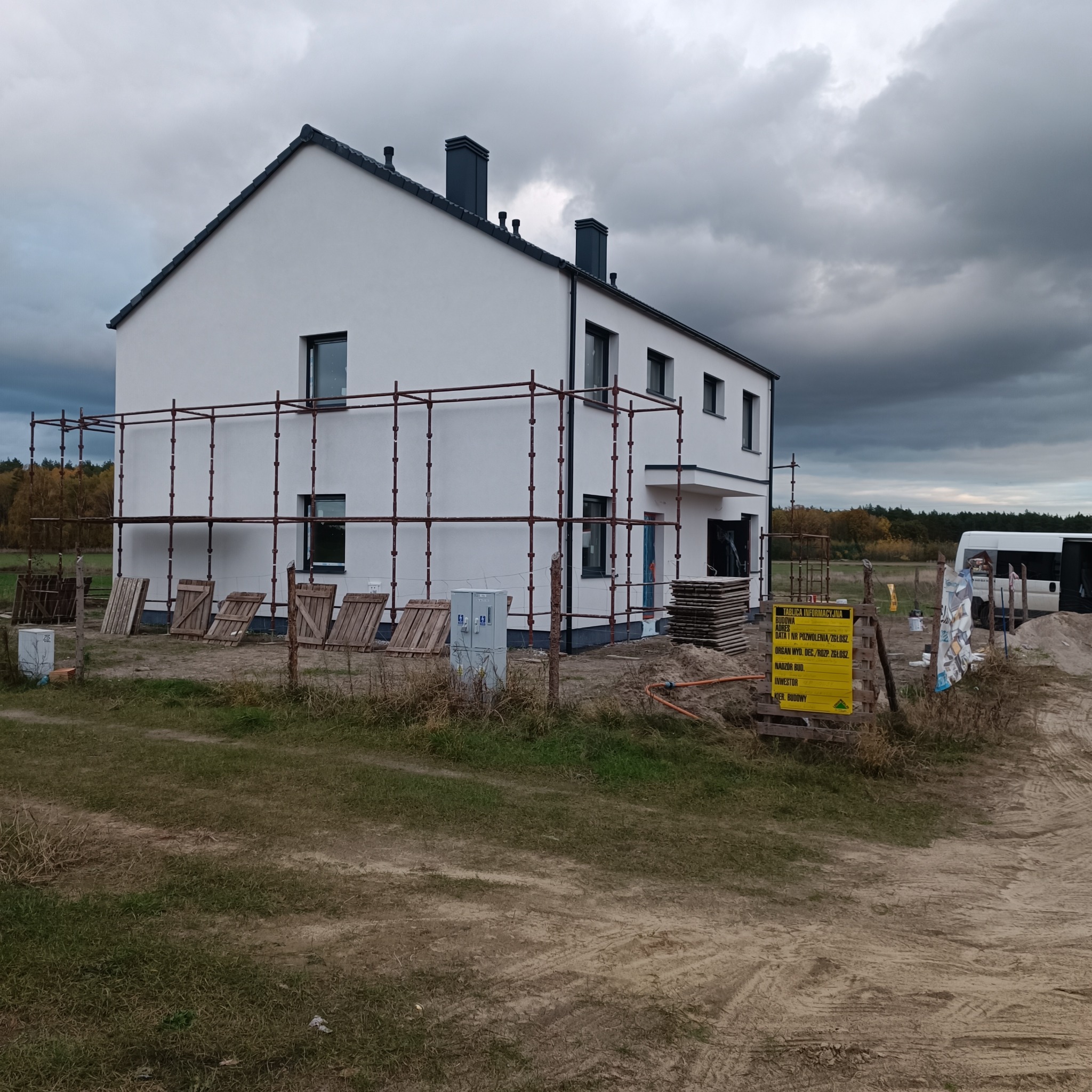 Nowoczesny, biały dom jednorodzinny w trakcie budowy, otoczony rusztowaniami, z widoczną tablicą informacyjną budowy i stertą materiałów budowlanych na zewnątrz, pochmurne niebo w tle.