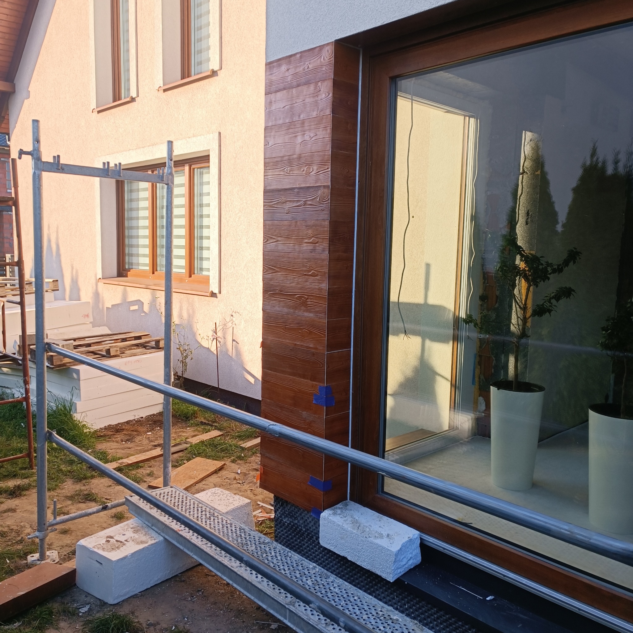 Wykończenie elewacji domu z drewnopodobną okładziną na narożniku, widoczne rusztowanie i materiały budowlane.