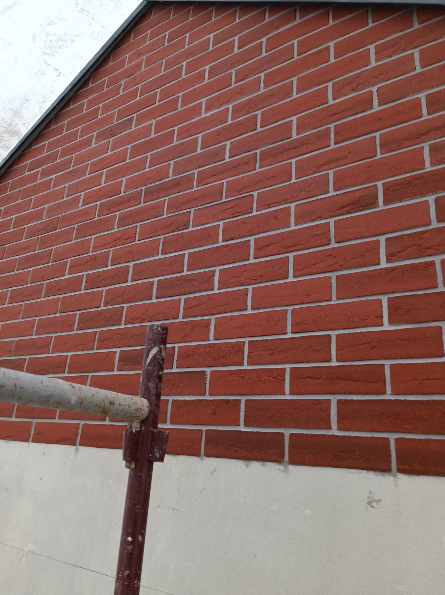 Fragment ściany zewnętrznej budynku z cegły klinkierowej, widoczne rusztowanie malowane na brązowo, podłoże pod cegłą szare.