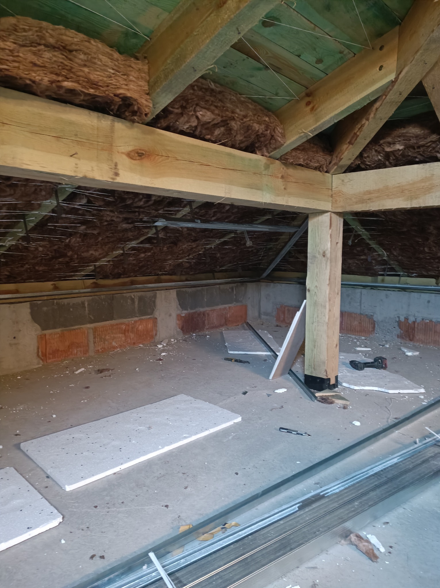 Poddasze w trakcie adaptacji: widoczne ocieplenie wełną mineralną pod zielonym dachem, murowane ściany, drewniane belki konstrukcyjne, profile metalowe i płyty karton-gips na podłodze, wiertarka...