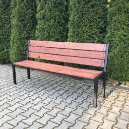 Usługi Stolarskie Konieczny - Ławka parkowa z drewnianym siedziskiem i oparciem w kolorze ciemnego orzecha oraz czarną metalową konstrukcją, ustawiona na kostce brukowej w Cielcach, na tle rzędu wysokich, zielonych tuj.