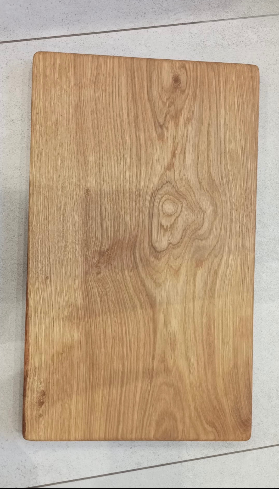 Prostokątna, dębowa deska do krojenia z widocznym usłojeniem drewna, leżąca na szarych płytkach podłogowych.